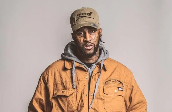 Man wearing a tan cap, gray hoodie, and tan jacket, with tattoos on face and neck, against a light gray background.