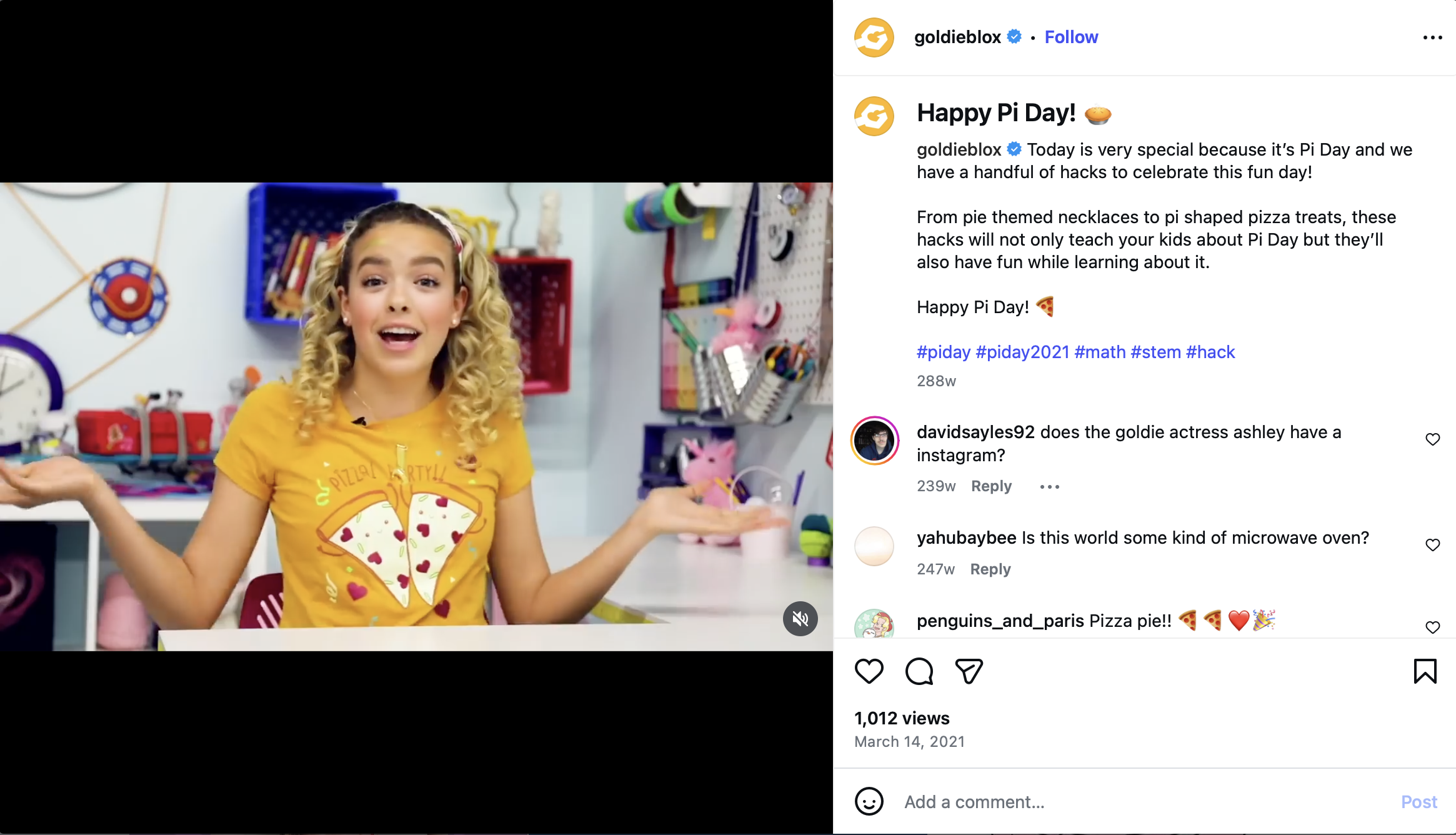 A young girl with curly blonde hair wearing a yellow t-shirt with pizza pieces on it, sitting in a craft room with colorful supplies on the wall behind her. She has an expressive face with raised eyebrows and her hands gesturing outward.