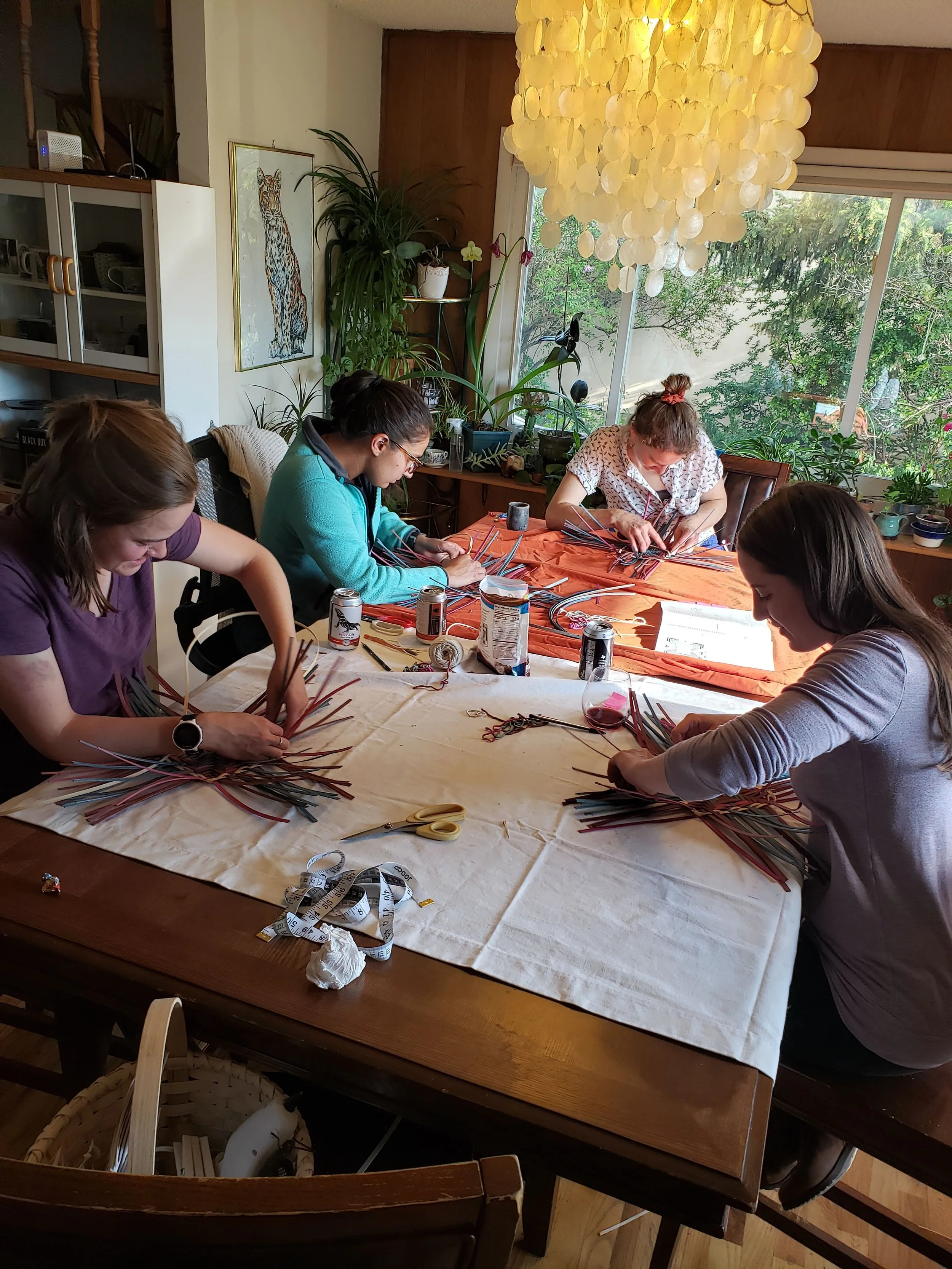 Four women sitting around a large table engaged in a craft activity involving colorful strips of material. The table is covered with fabric, scissors, tape measure, and cans. The room has large windows with greenery outside, and various plants and artwork decorate the space. A chandelier made of circular discs hangs overhead.