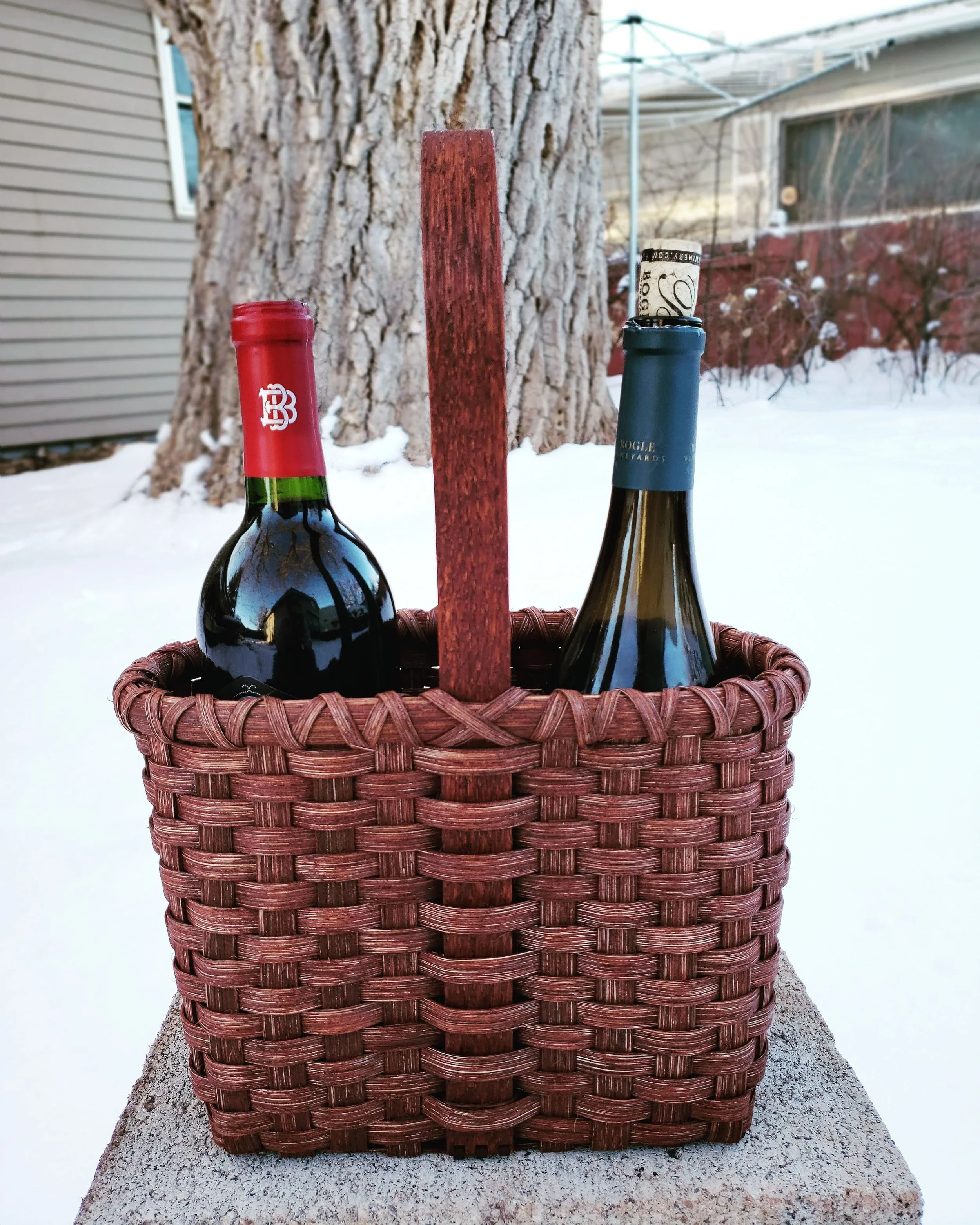 A wicker basket with a wooden handle, containing two bottles of wine, is placed on a concrete surface outdoors. One bottle has a red foil and the other has a blue foil. Snow and a large tree are visible in the background.
