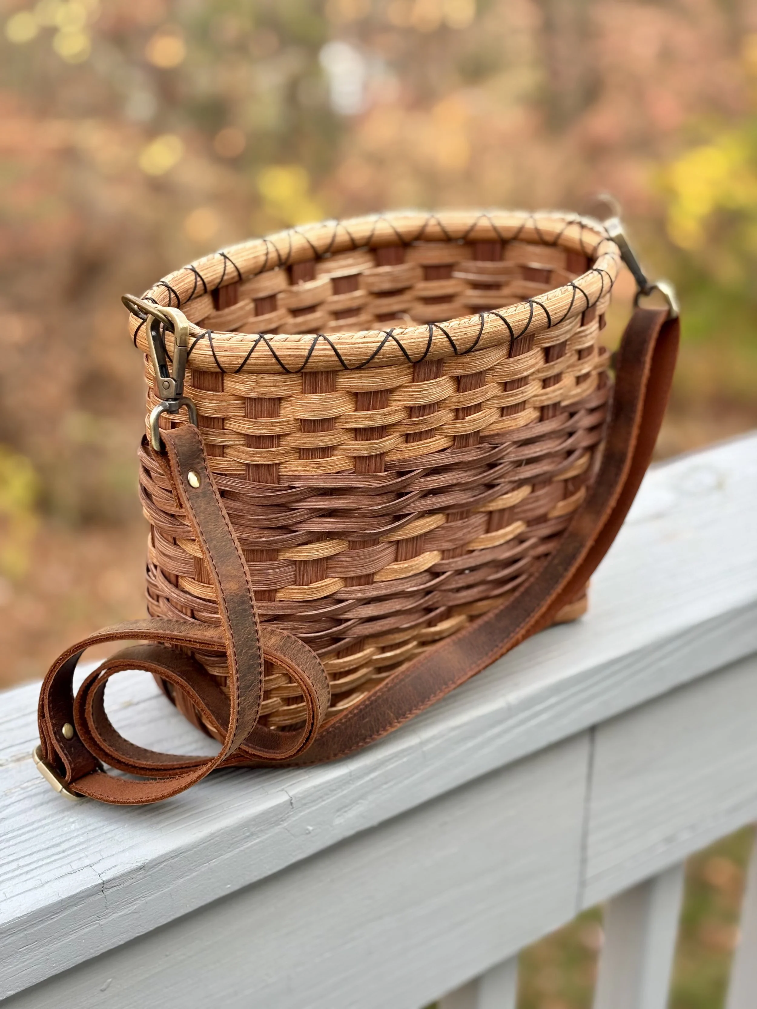 A woven wicker basket with leather straps and brass buckles, sitting on a white wooden surface outdoors, with blurred autumnal trees in the background.
