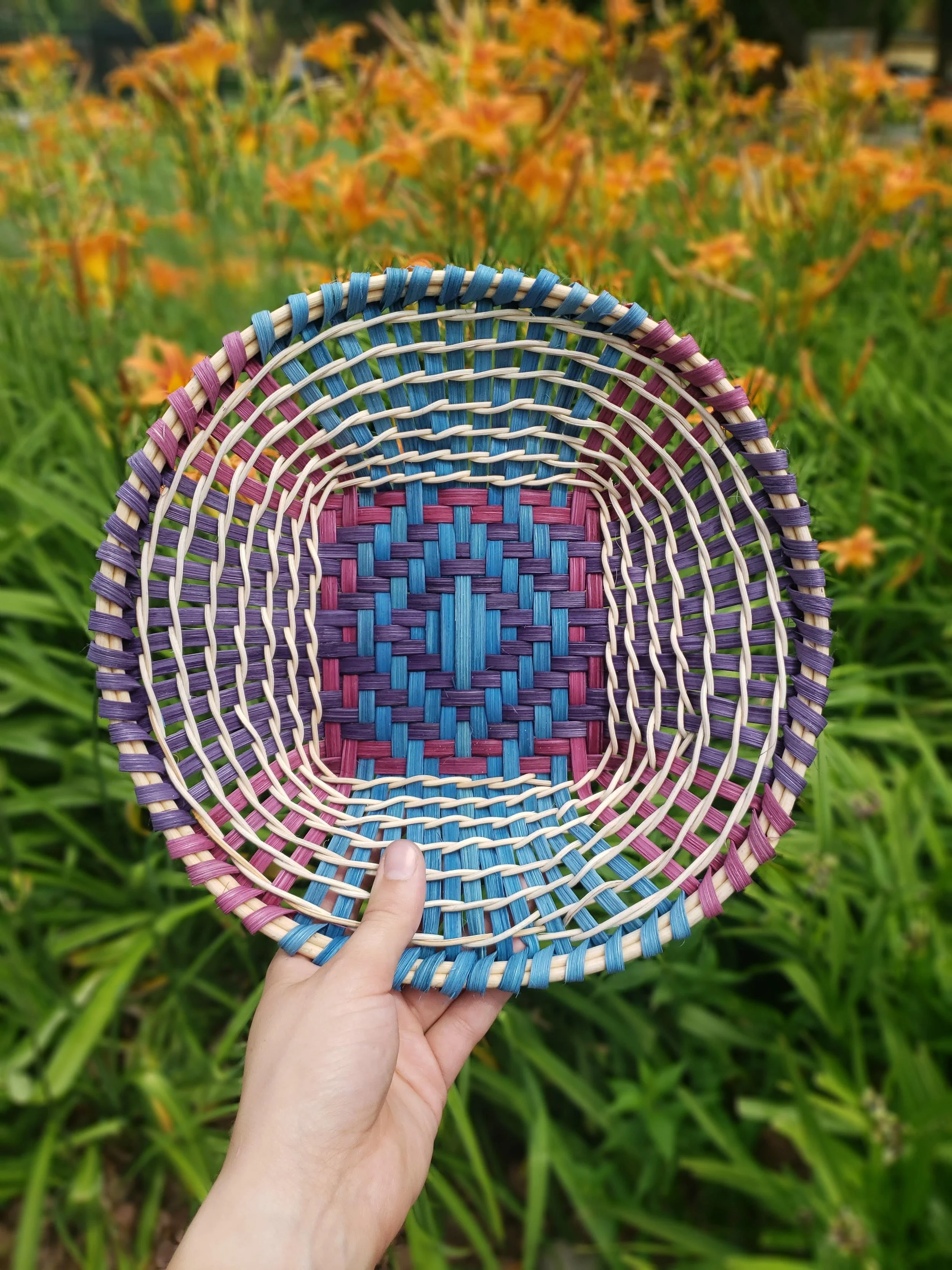 Close-up of a hand holding a colorful woven basket with pink, purple, blue, and beige strands, against a blurred background of green foliage and orange flowers.