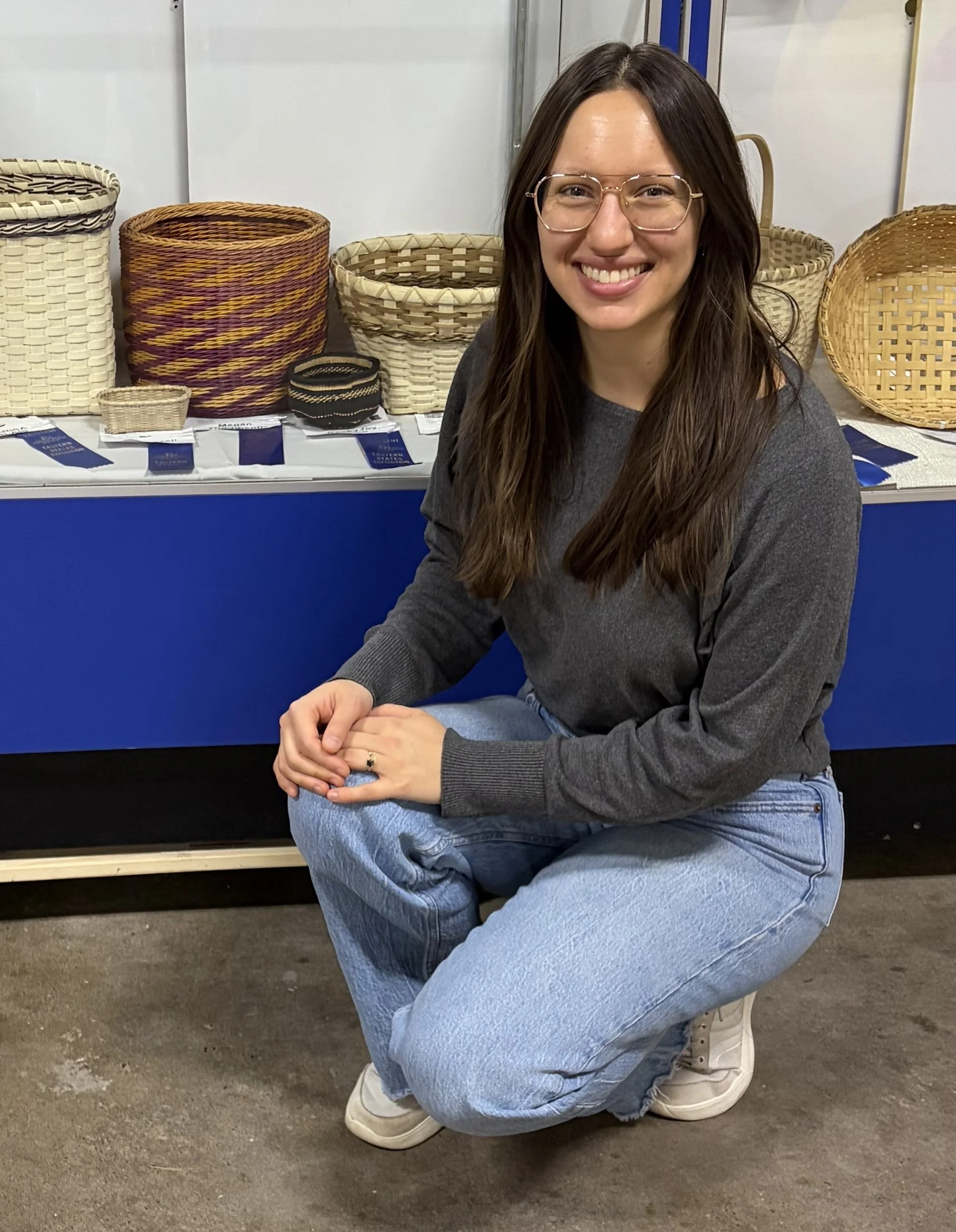 A woman with long brown hair, wearing glasses, a gray sweater, and blue jeans, crouching on the floor in front of woven baskets at an indoor market or craft fair, smiling at the camera.