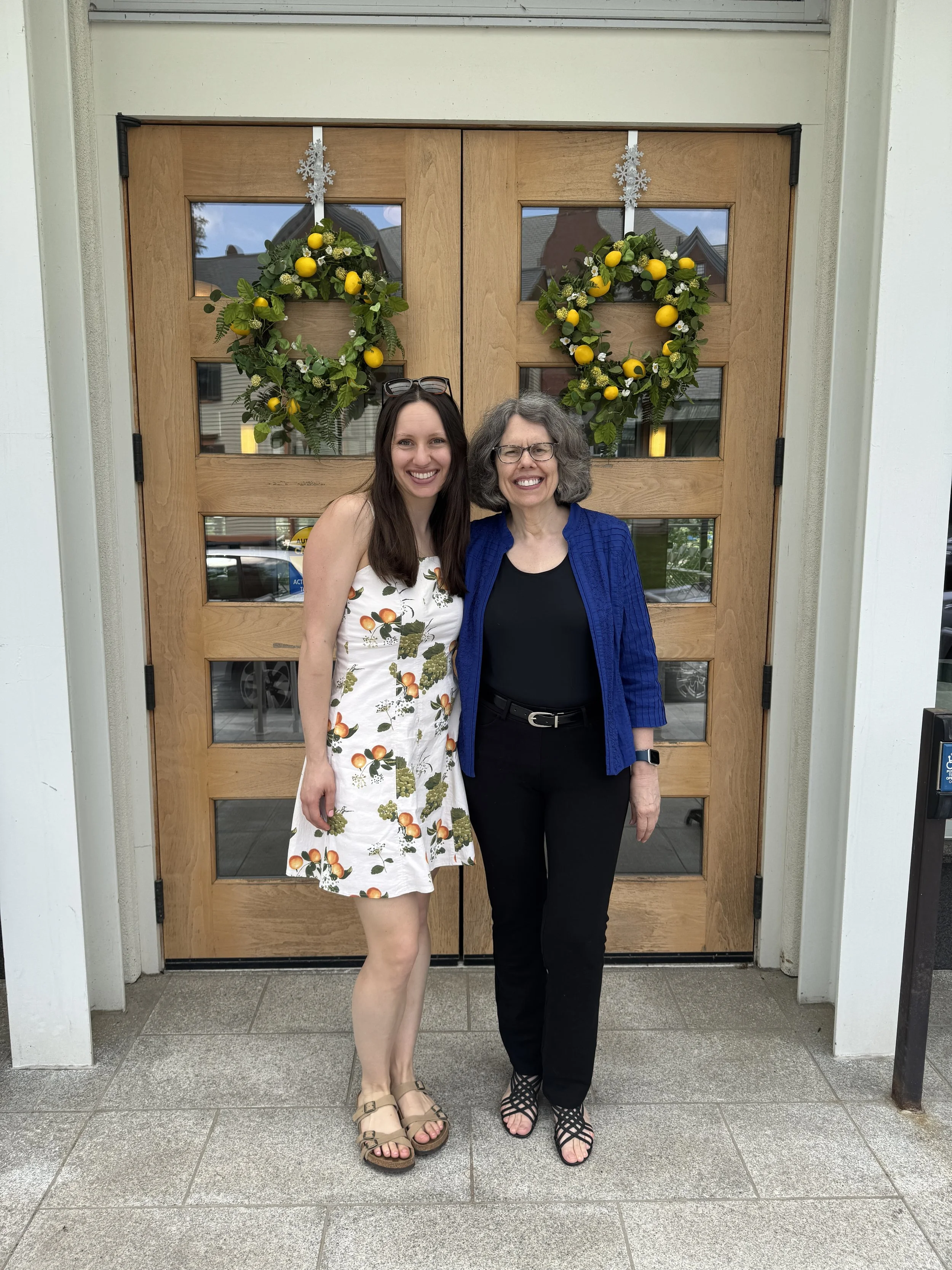 Two women standing in front of a wooden door with holiday wreaths and snowflake decorations, smiling at the camera.