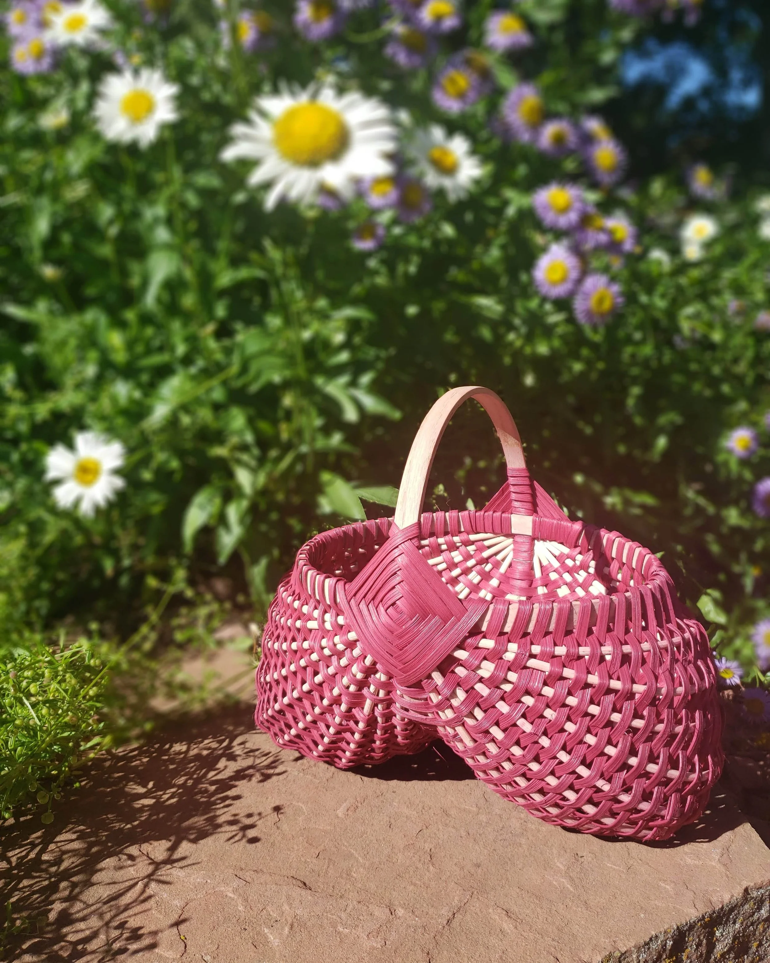 Pink woven basket on a stone surface with shadow of the basket casts on the ground, surrounded by green plants and purple and white flowers in the background.