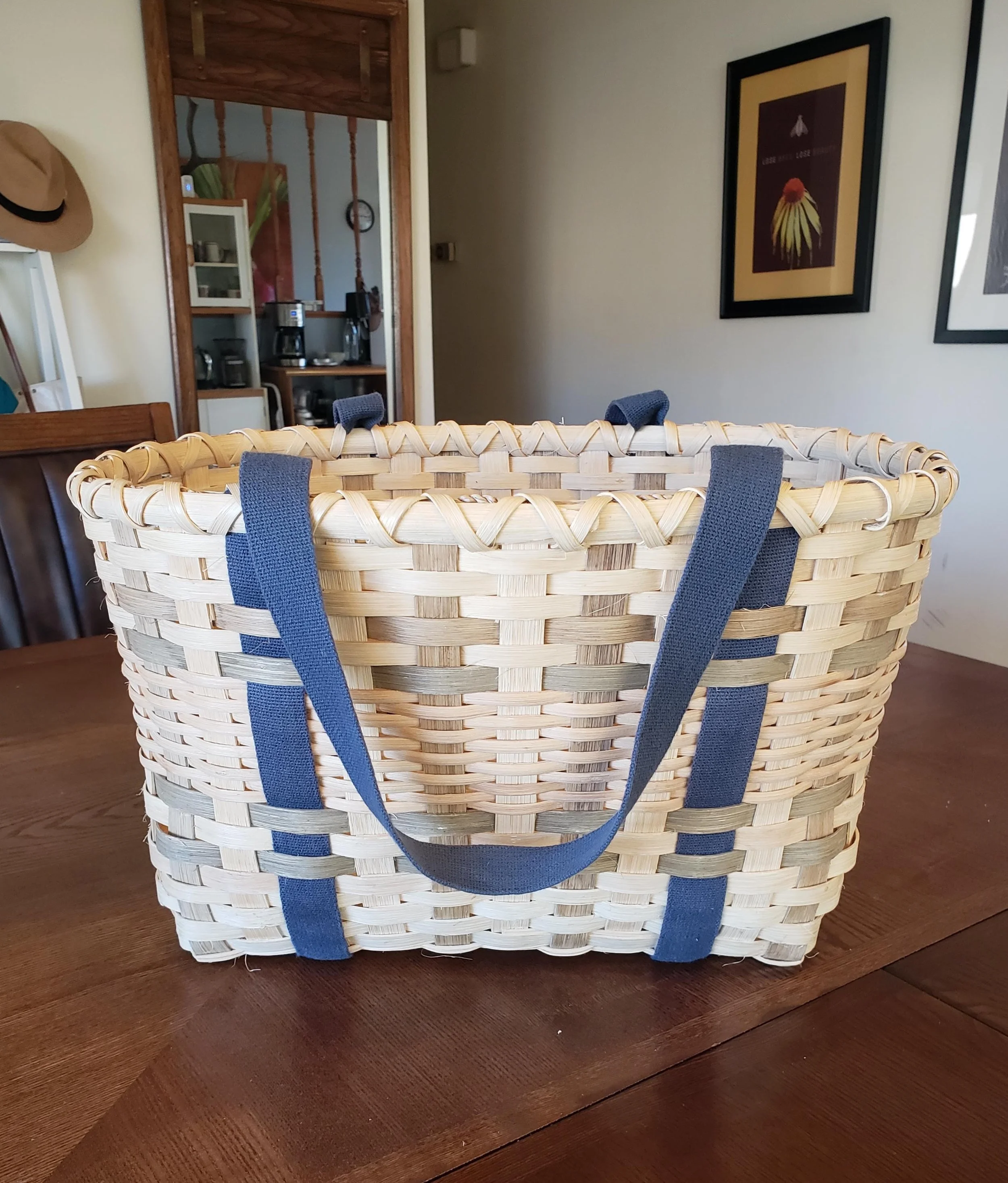 Woven basket with blue straps placed on a wooden surface in a room with framed artworks on the wall.