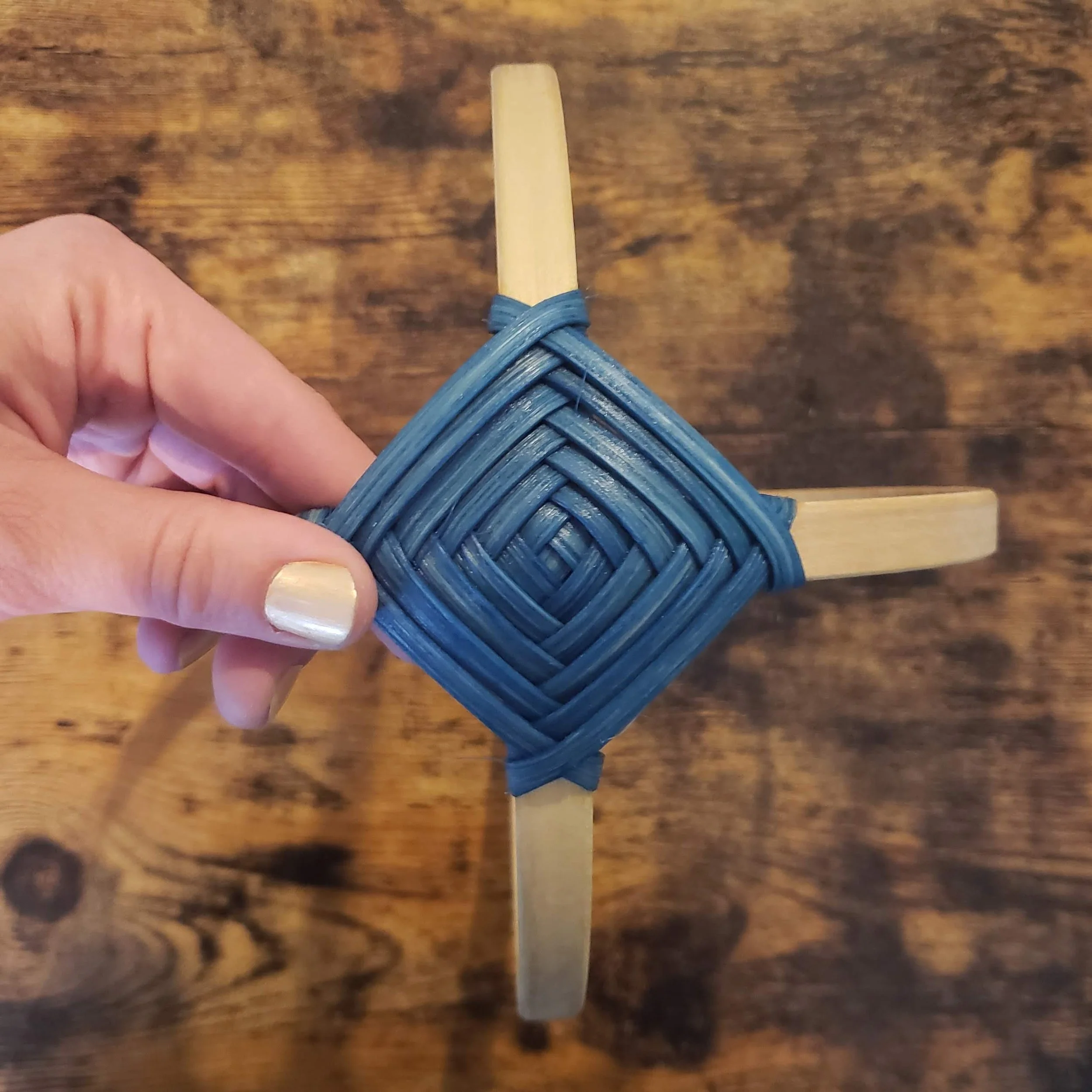 Close-up of a hand holding a woven blue item with a wooden stick protruding from it on a wooden surface.