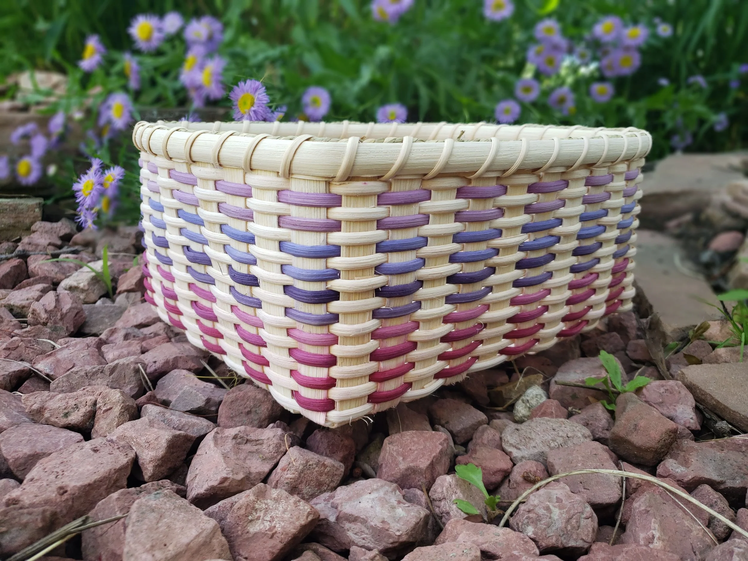 A woven basket with purple, pink, and cream stripes, placed on reddish gravel, with purple flowers and green foliage in the background.