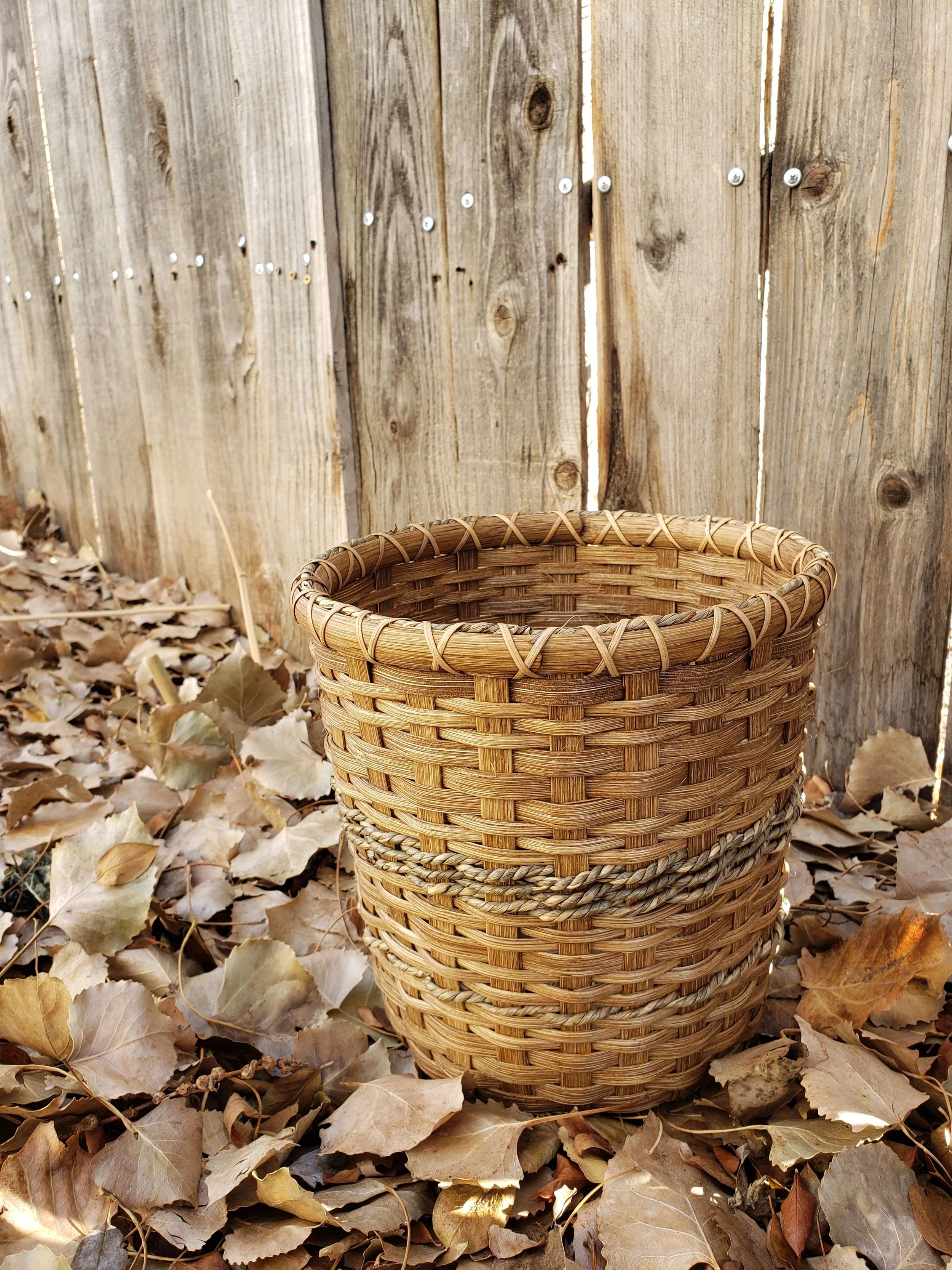 A woven basket resting on fallen dry leaves against a wooden fence.