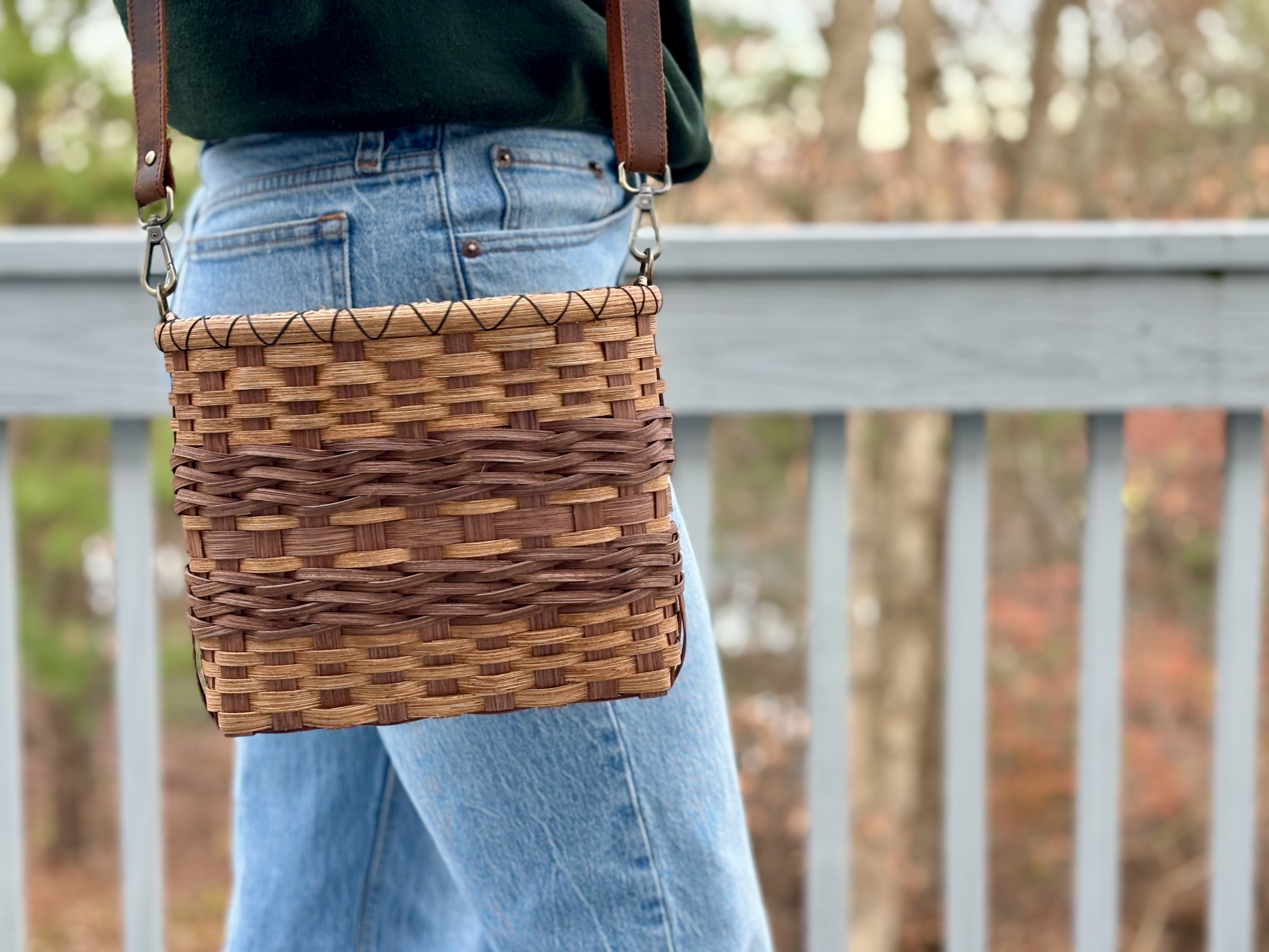 Person wearing blue jeans and a black jacket carrying a small, woven brown basket with a leather strap on a walk outdoors with trees in the background.