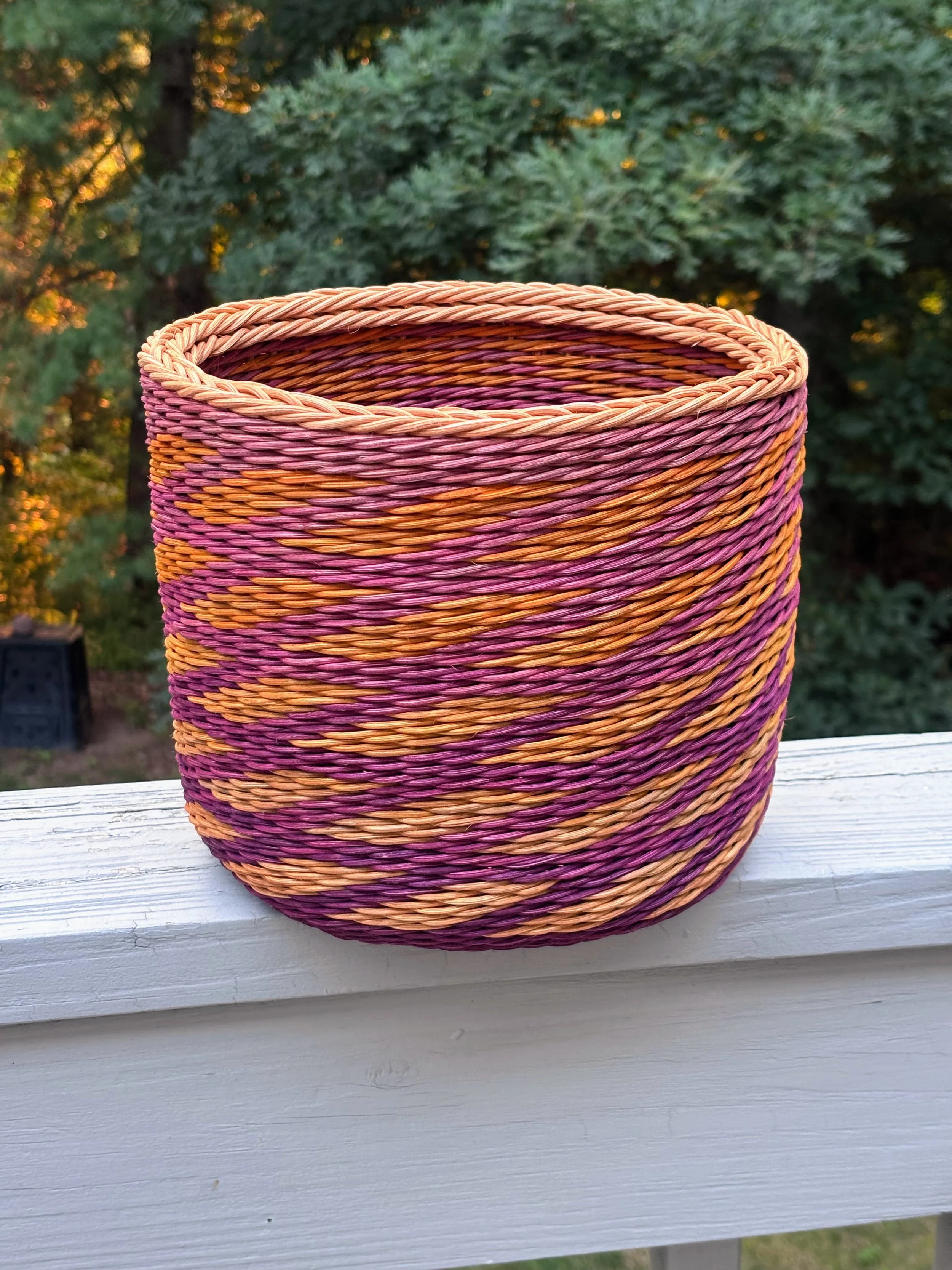 A colorful woven basket with pink, orange, and purple hues, placed on a white wooden railing outdoors with a backdrop of green trees.