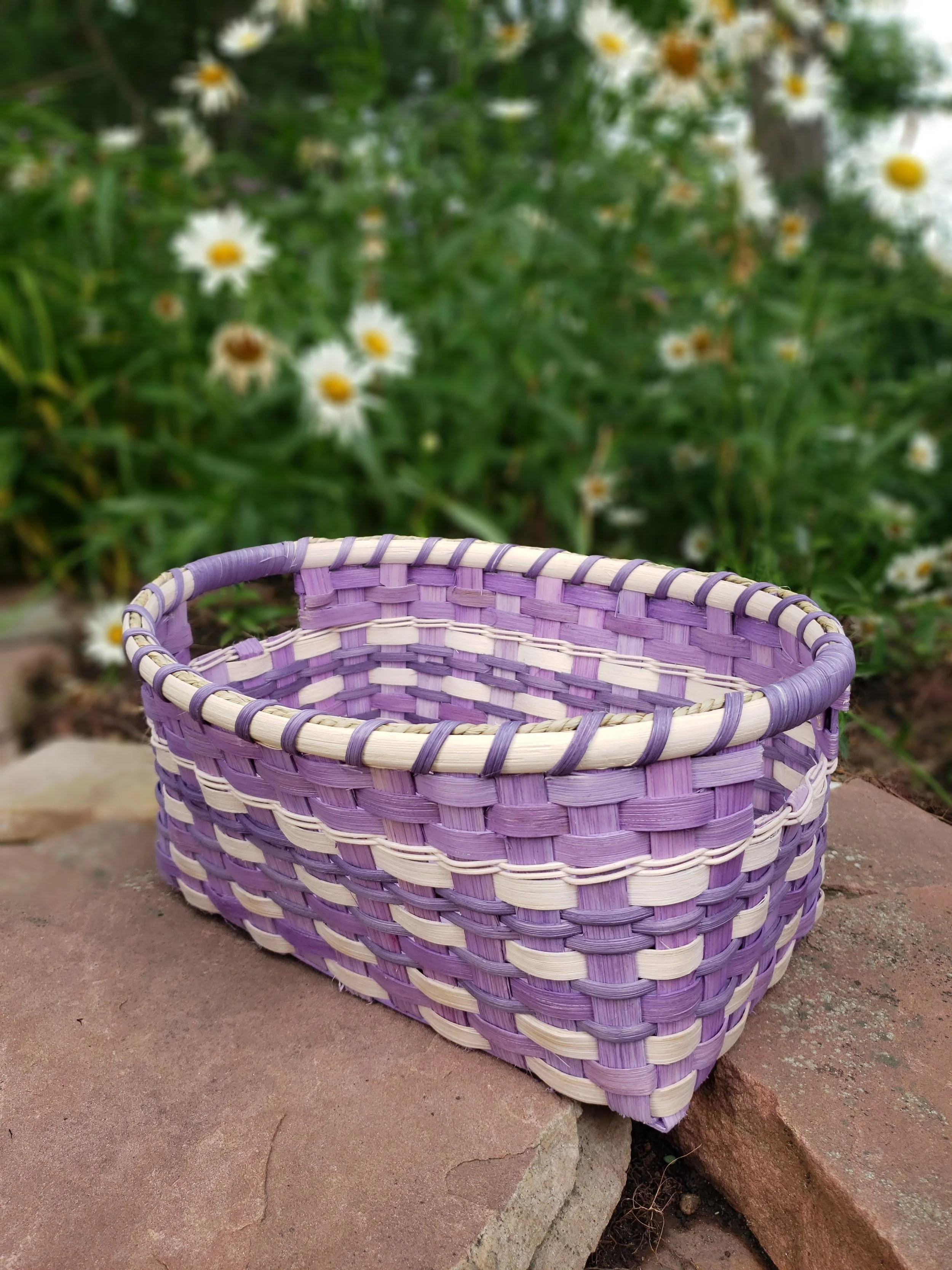 A purple and white woven basket placed on a concrete surface outdoors, with green foliage and white daisies in the background.
