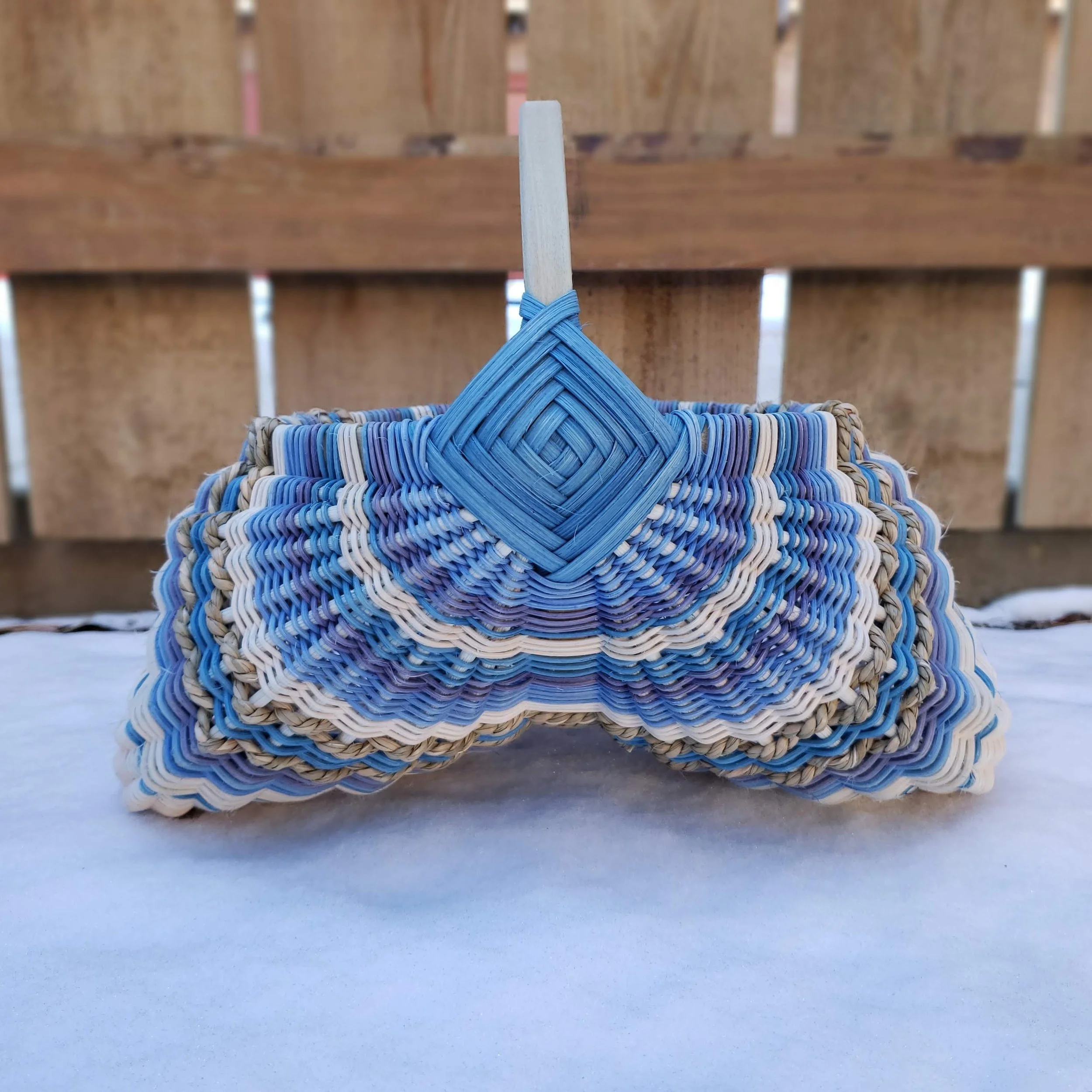 Close-up of a woven basket hanging from a string tied to a wooden stick, with a background of a wooden fence and snow on the ground.