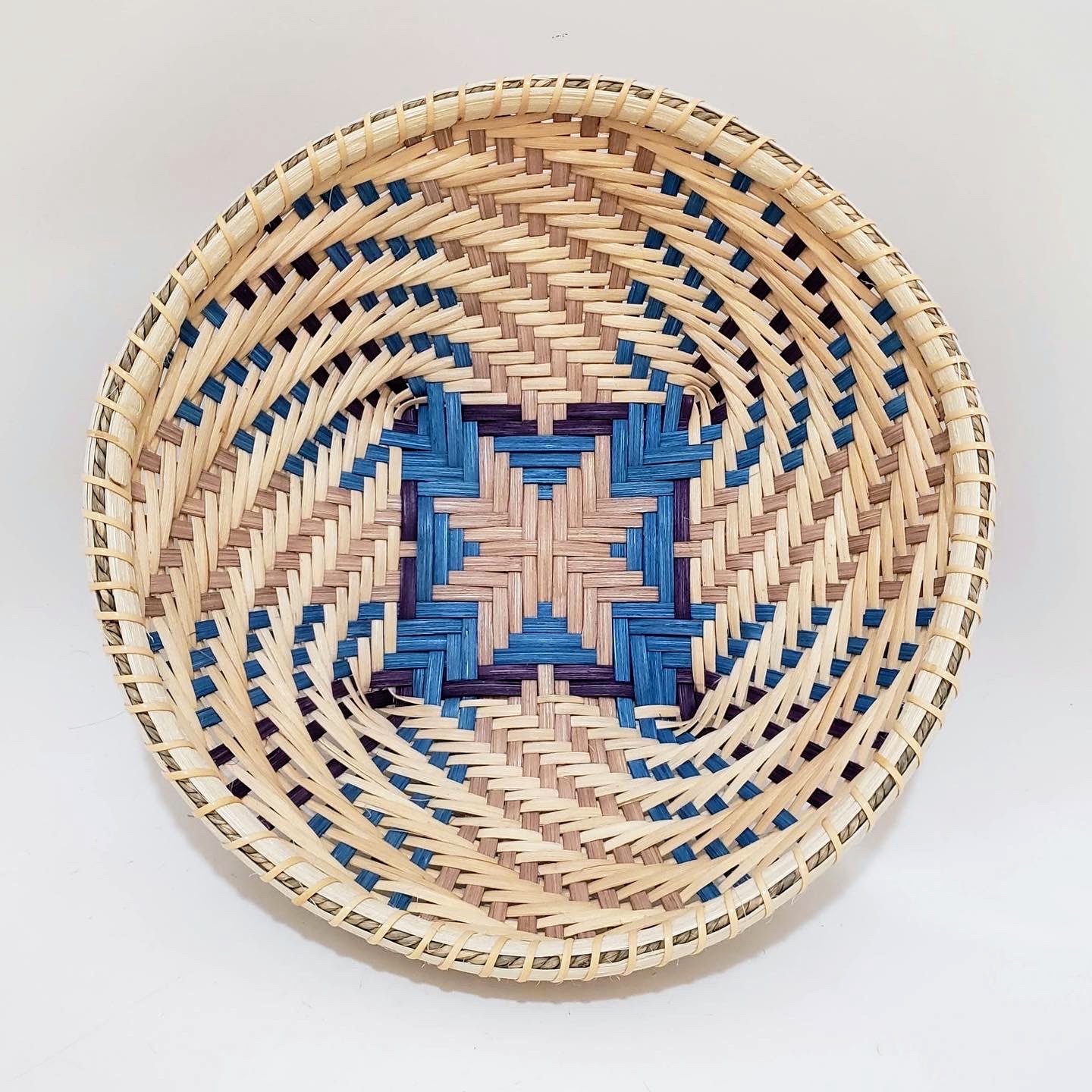 Round woven basket with a geometric pattern in blue, pink, and natural straw colors, on a white surface.