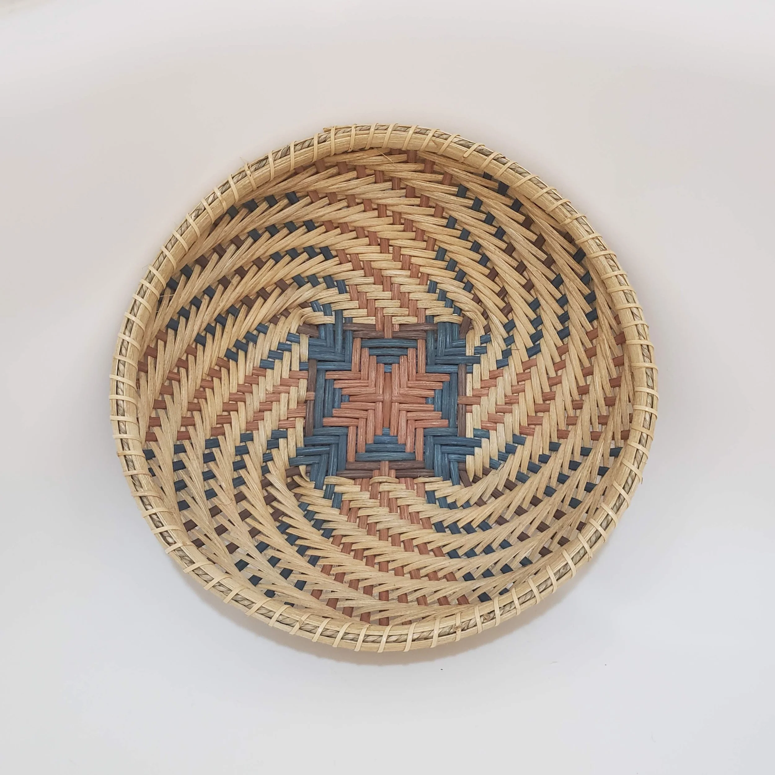 A woven basket with a circular pattern, made of natural and colored straw, placed on a white background.