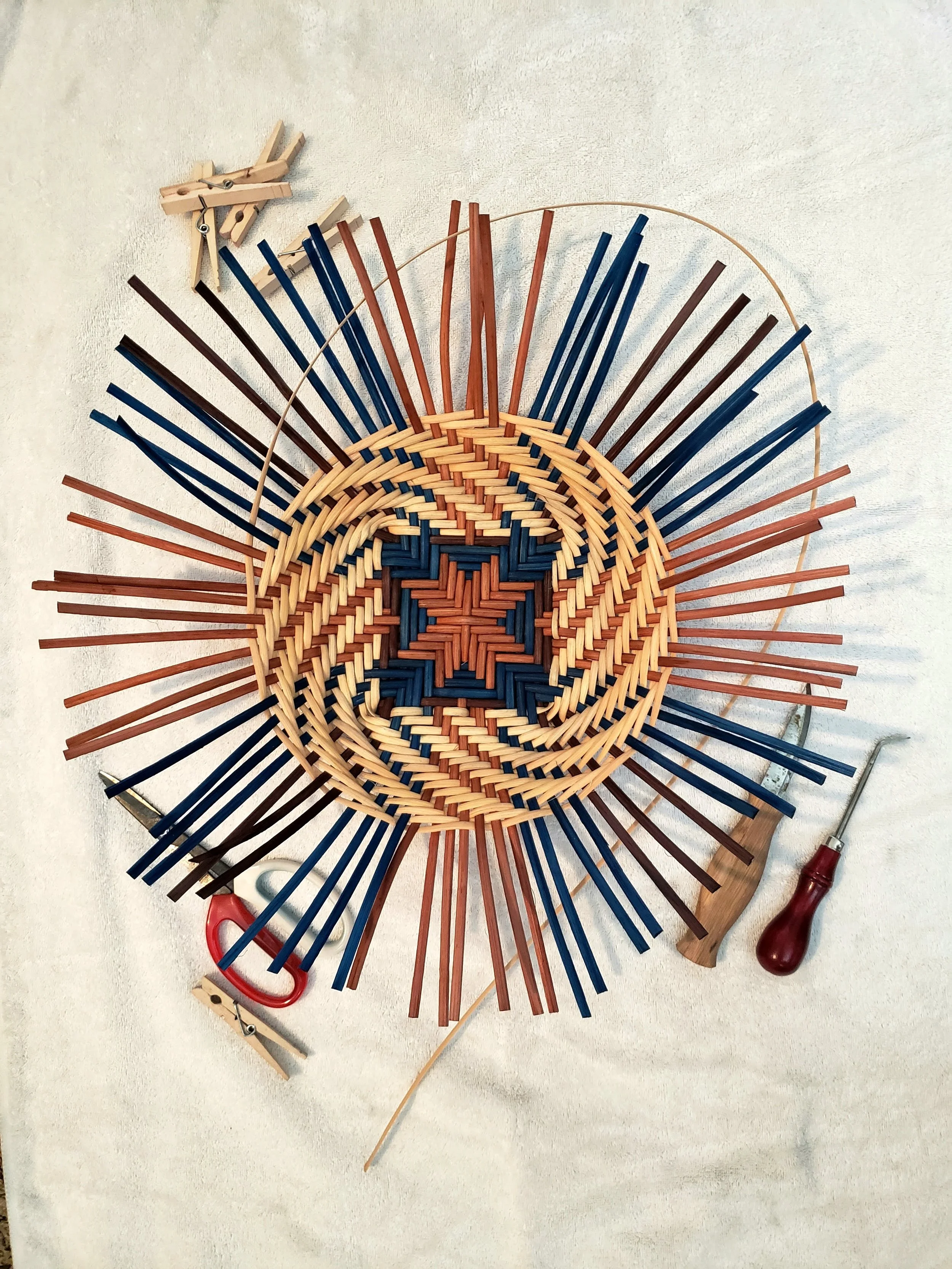 Unfinished woven basket with a circular pattern, surrounded by tools including clothespins, scissors, a small screwdriver, and a piece of wire, on a white surface.