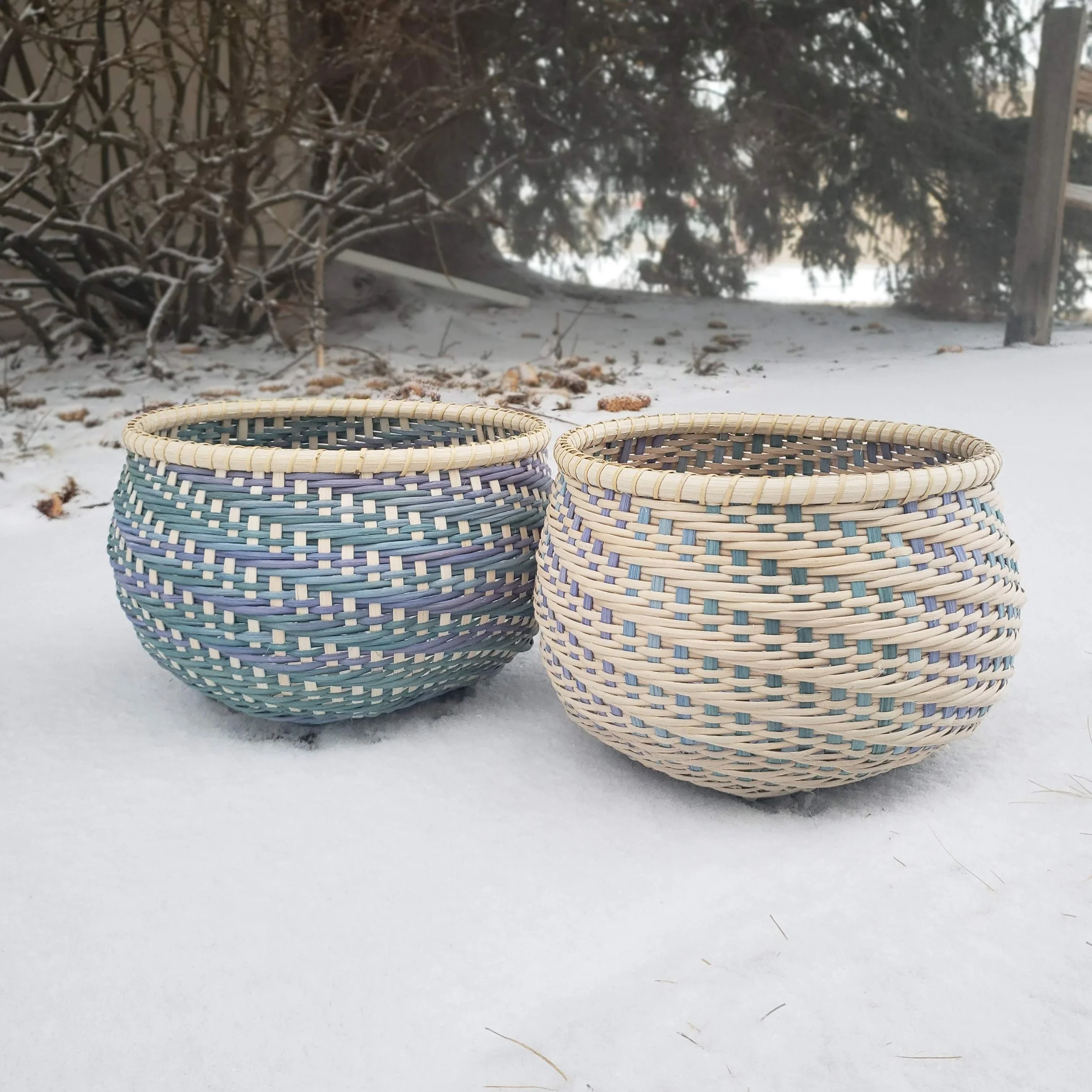 Three woven baskets in front of snowy ground with trees and wooden fence in the background.