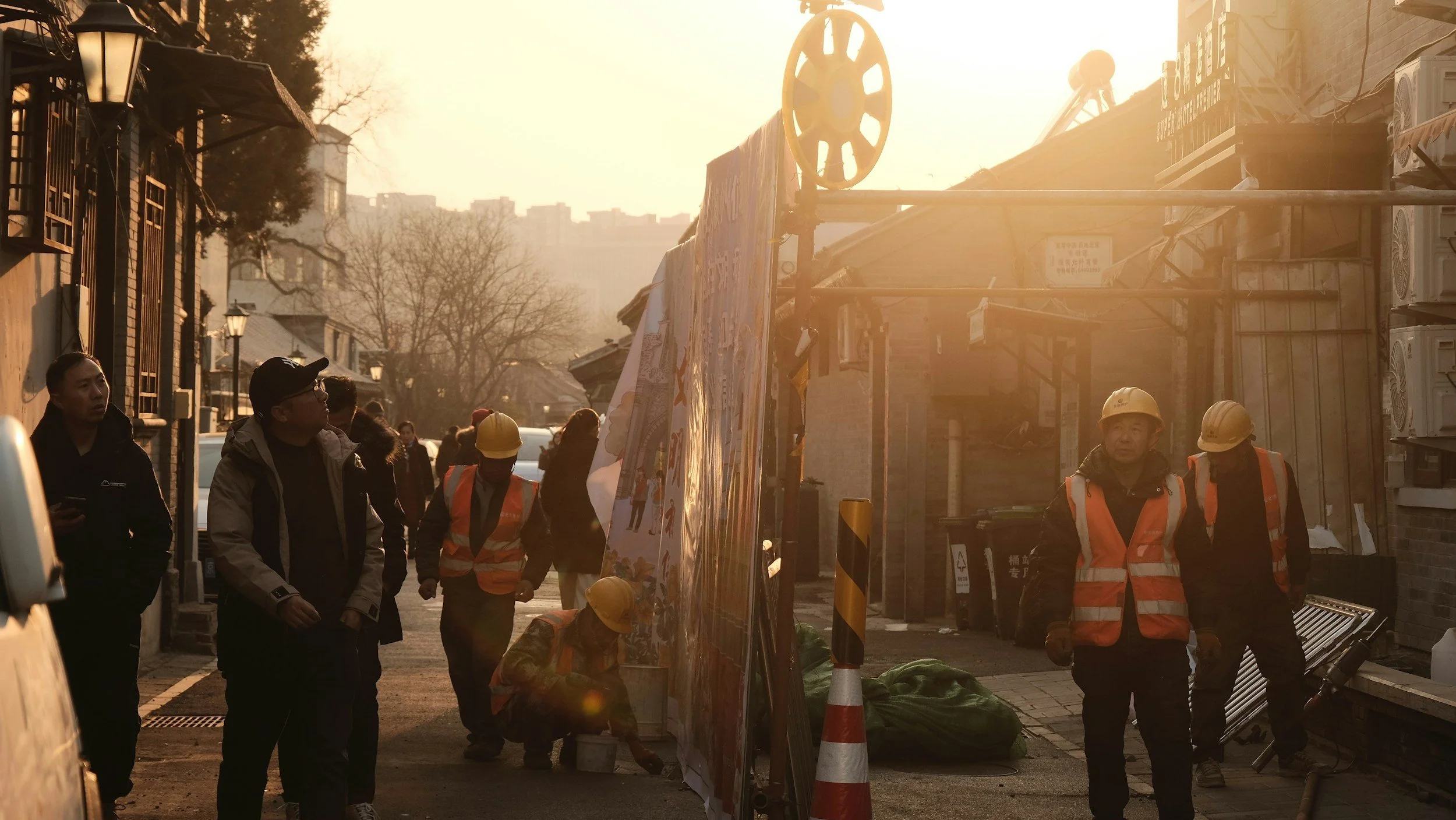 Operai con caschi e giubbotti arancioni lavorano in una strada in una zona urbana al tramonto, probabilmente durante lavori di costruzione o riparazione.