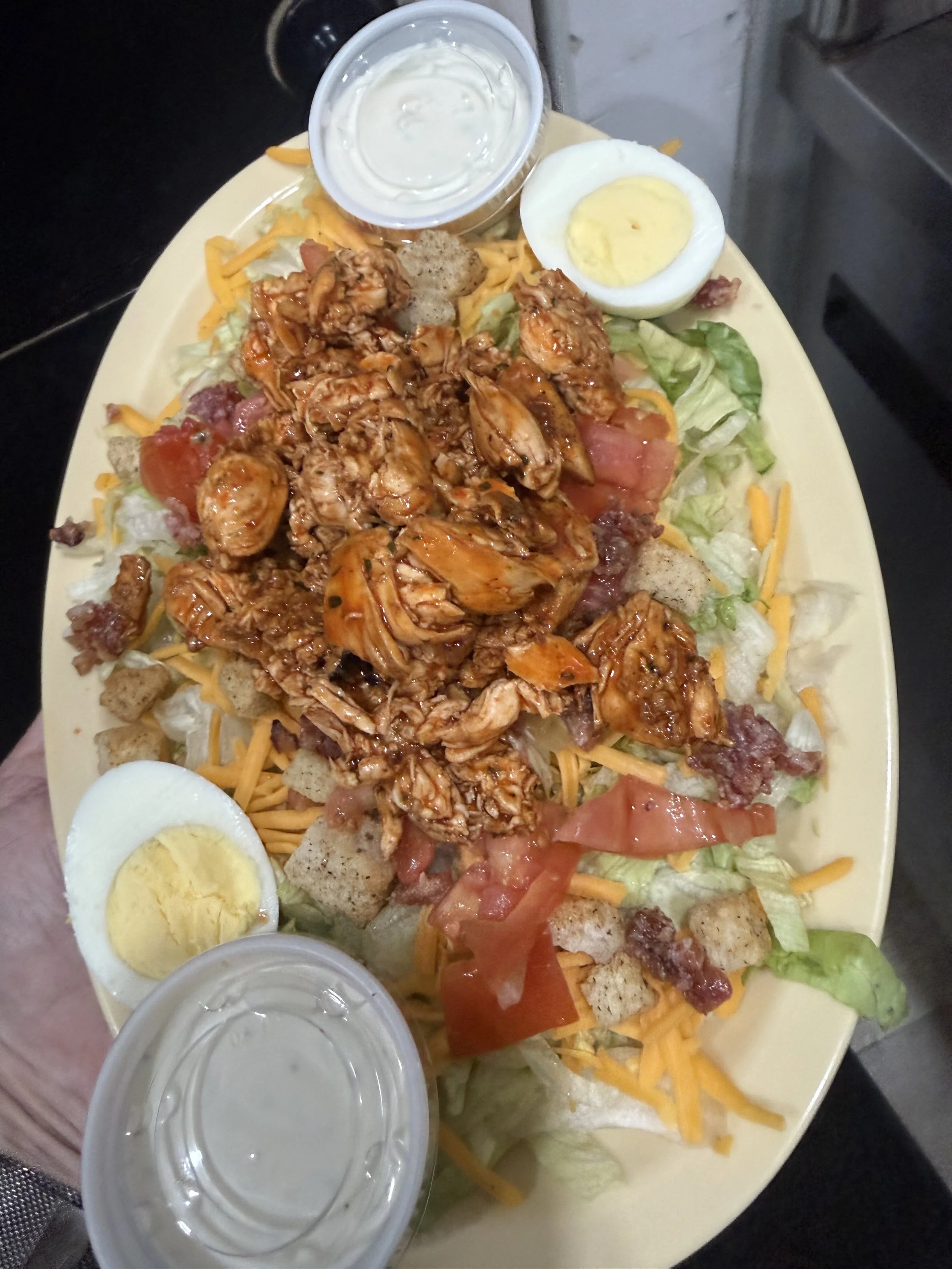 A plate of salad topped with grilled chicken, shredded cheese, croutons, chopped tomatoes, and lettuce, accompanied by sliced boiled eggs, a cup of ranch dressing, and a glass of water.
