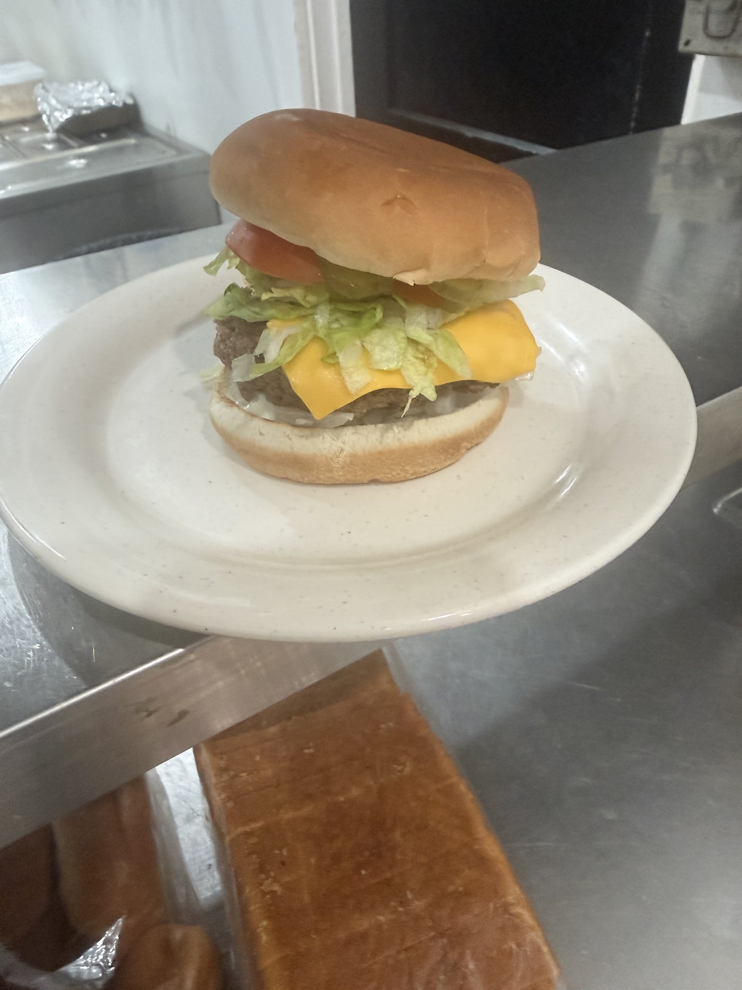 A cheeseburger with lettuce, tomato, and a beef patty on a round sandwich bun, served on a white plate on a metal surface.