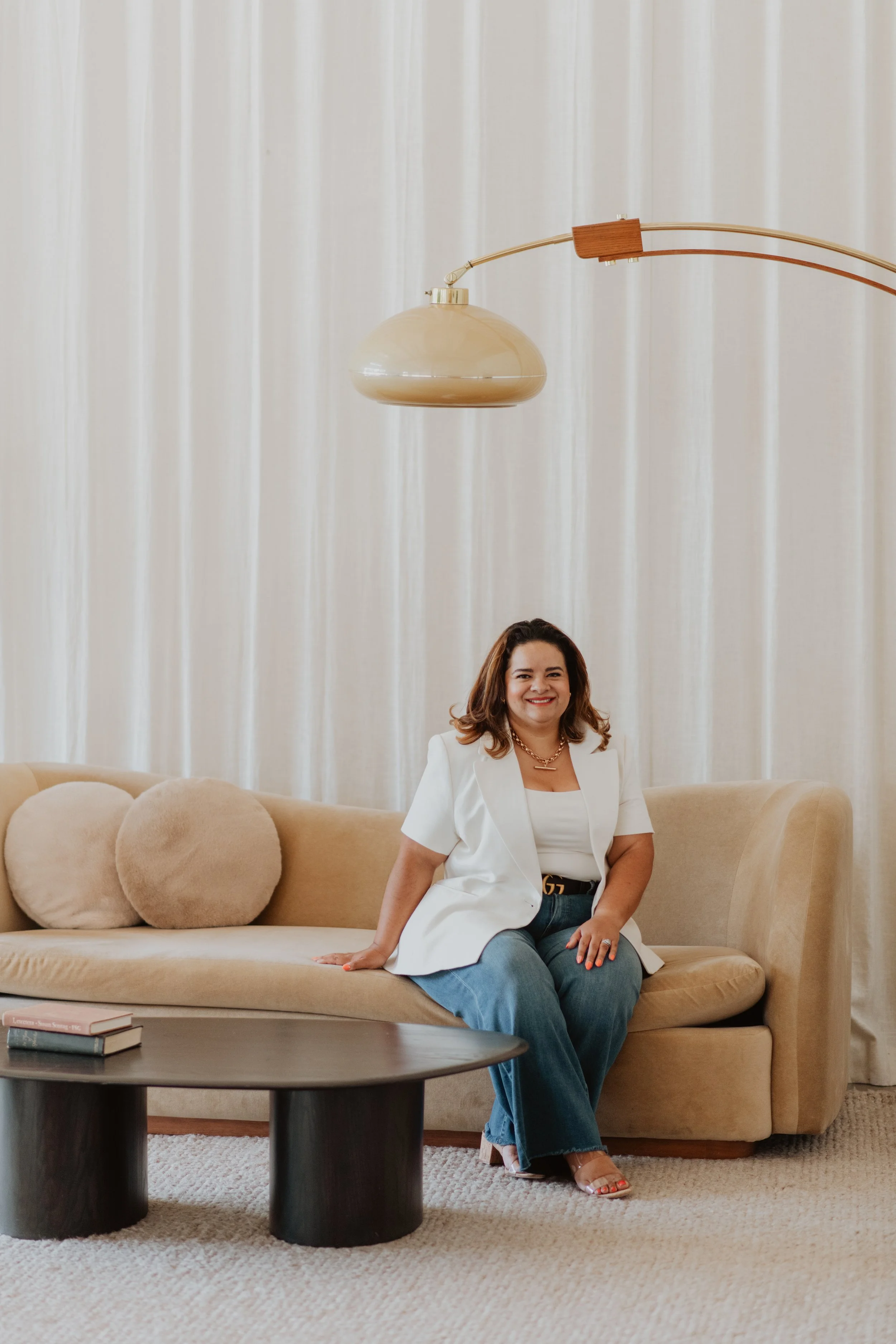 A woman sitting on a beige curved sofa with plush round pillows, wearing a white blazer, white top, and blue jeans, smiling at the camera in a living room with beige carpet and cream curtains.