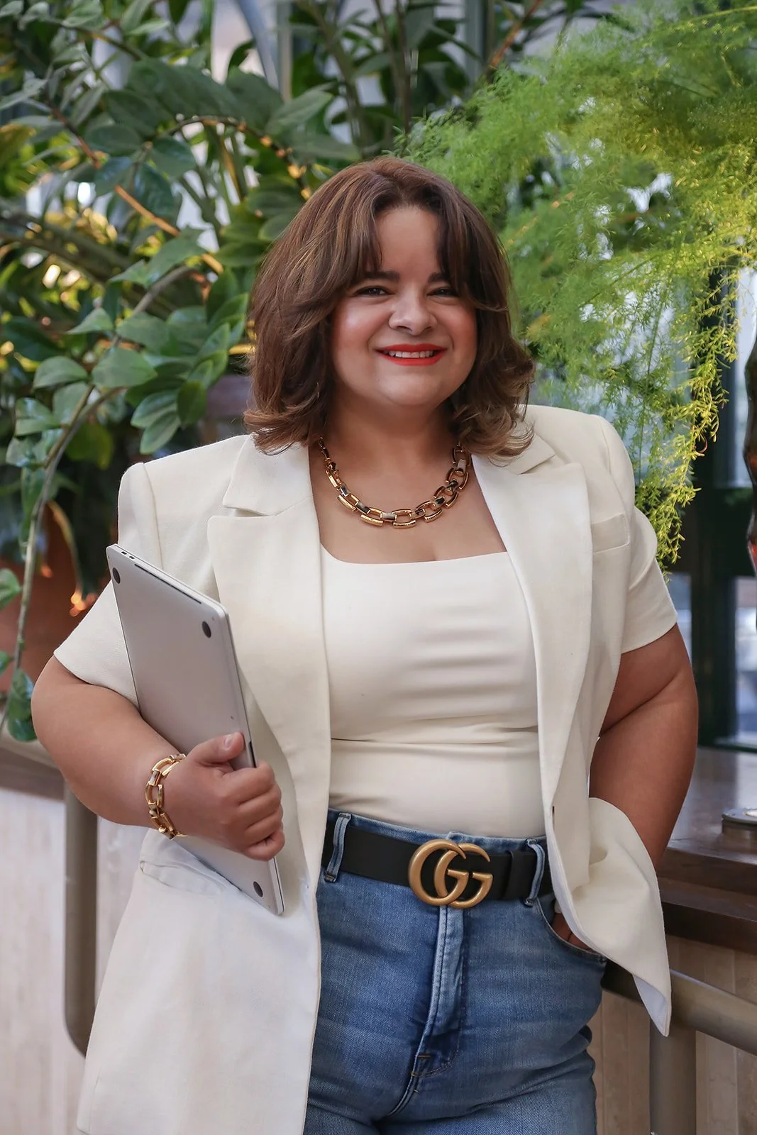 A woman with shoulder-length brown hair smiling, dressed in a cream blazer, matching top, and blue jeans, standing in front of green foliage, holding a laptop.