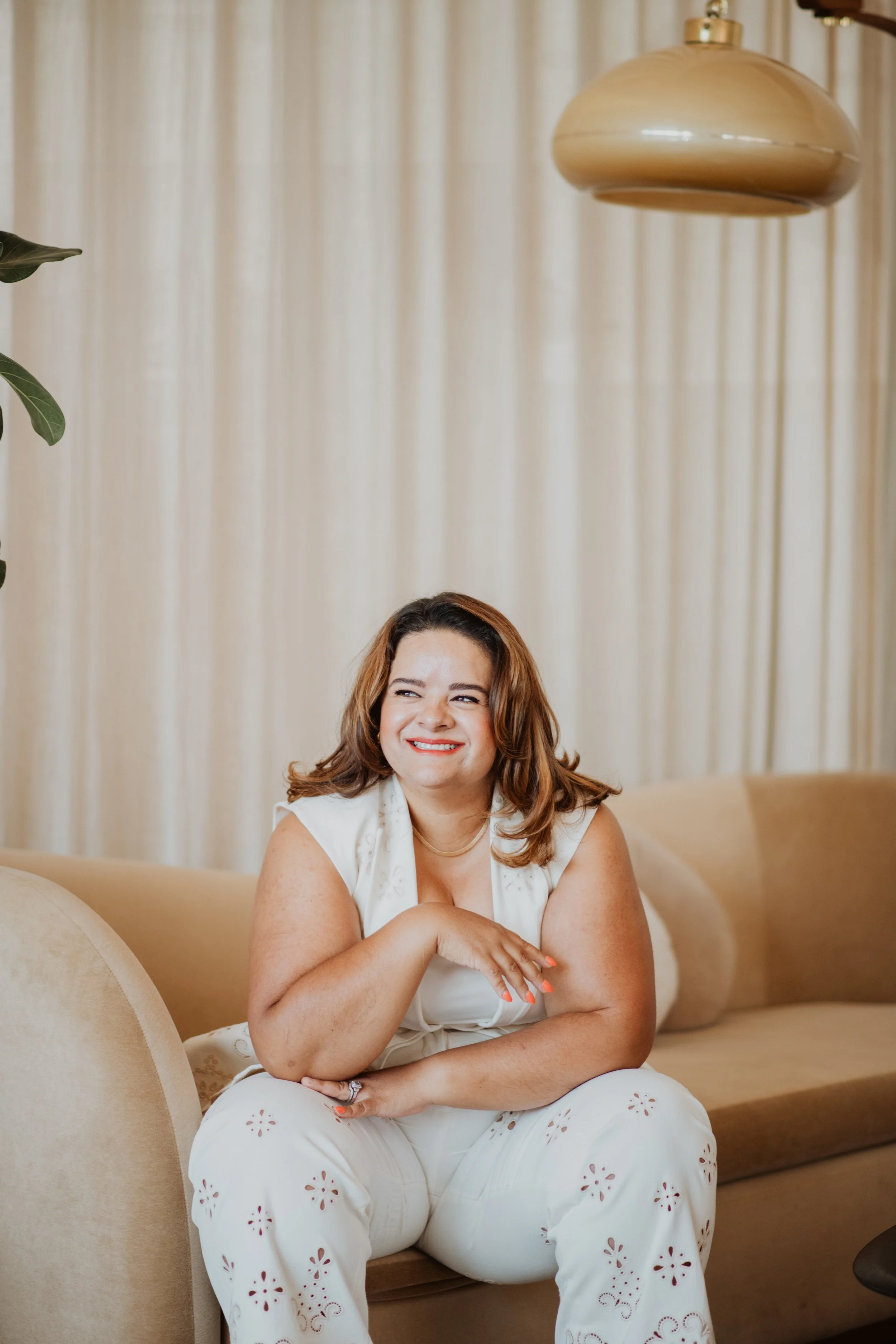 A woman with light skin and brown hair sitting on a beige sofa in a room with cream-colored curtains. She is smiling, wearing a white sleeveless top and white pants with cut-out floral patterns, and has her arms crossed over her lap.
