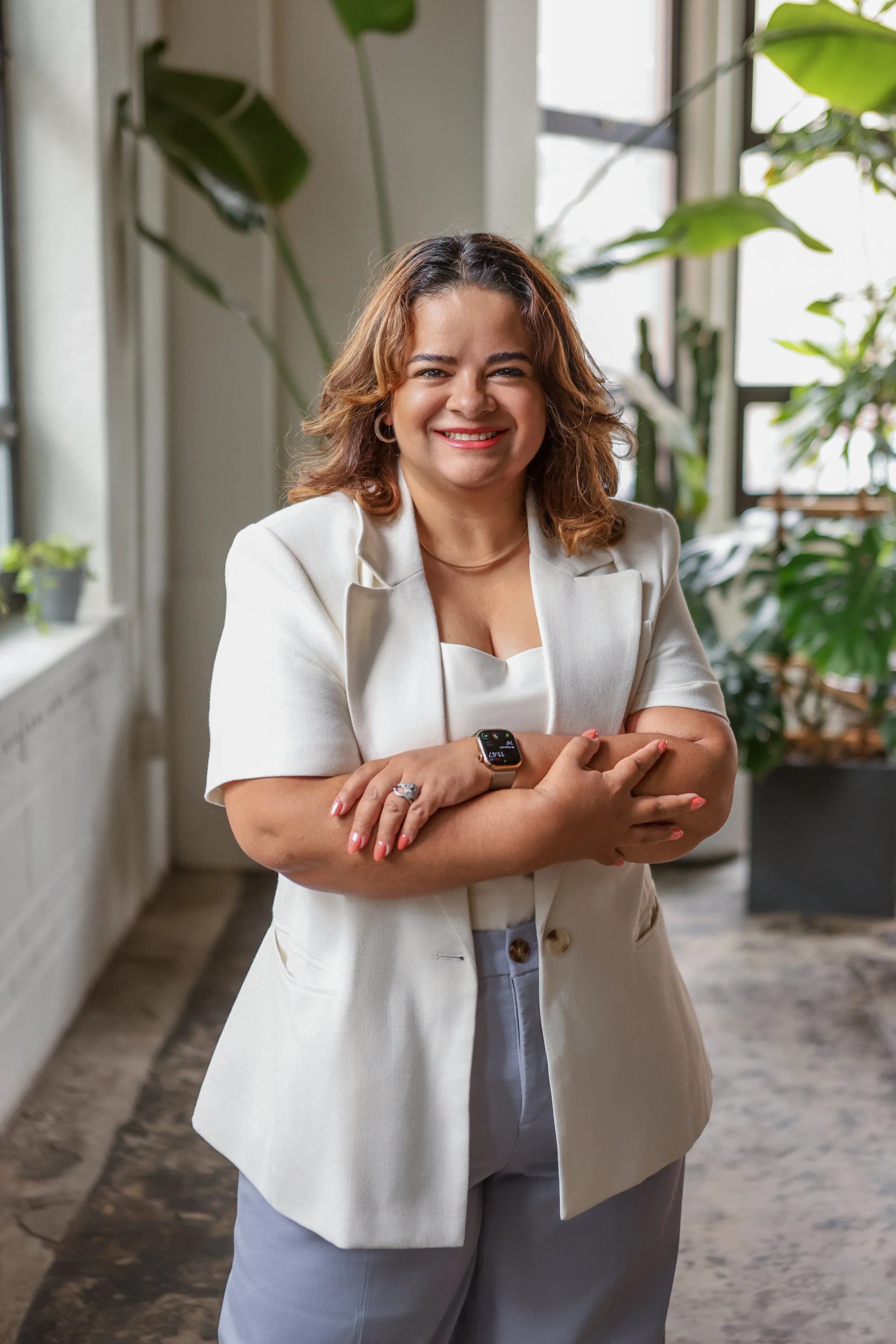 A woman with shoulder-length curly hair, wearing a cream blazer over a white top, standing in a room with large windows and green plants, smiling with arms crossed, and wearing a smartwatch.
