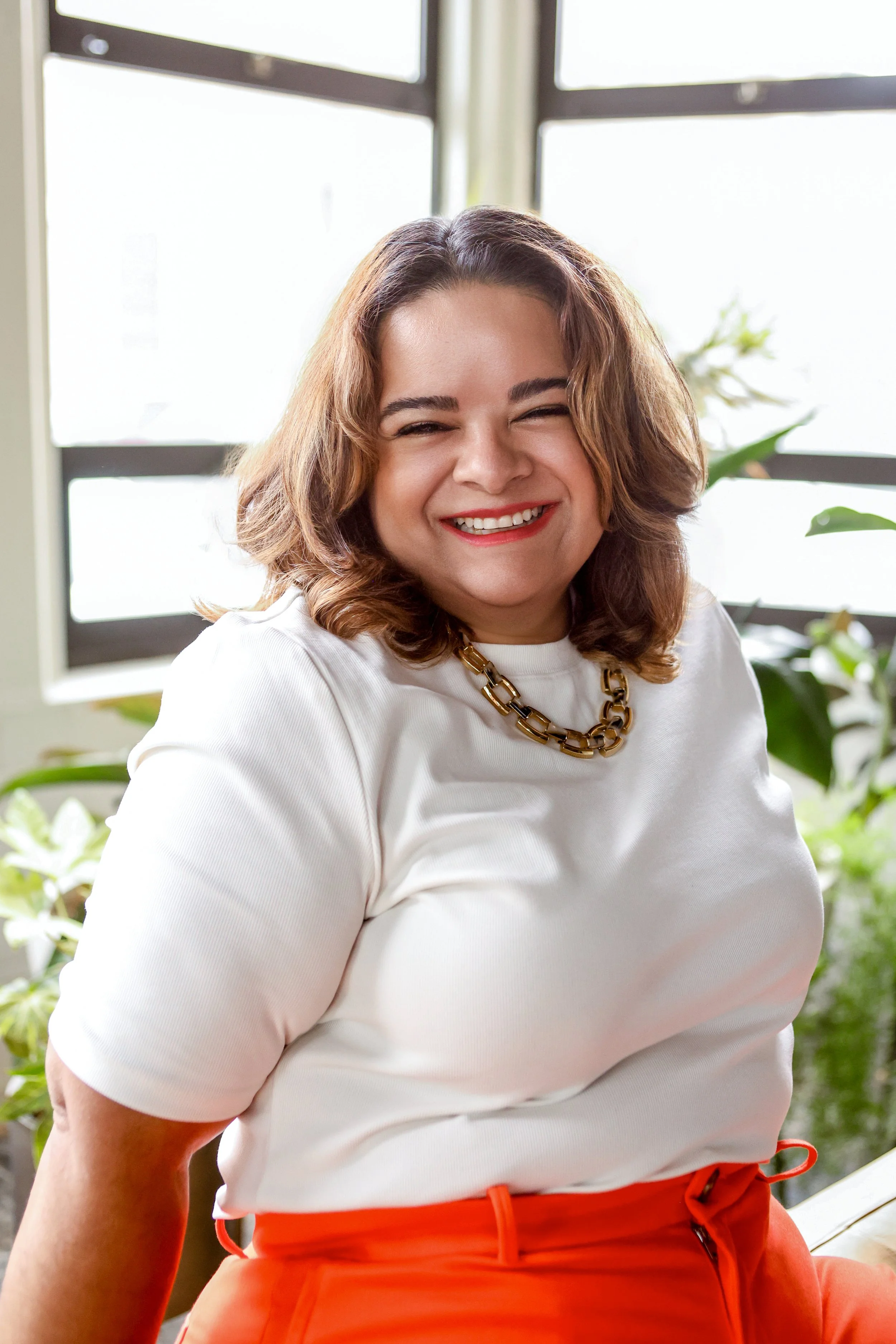 A woman with shoulder-length brown hair, smiling, wearing a white top and orange pants, in a bright indoor space with plants and large windows.