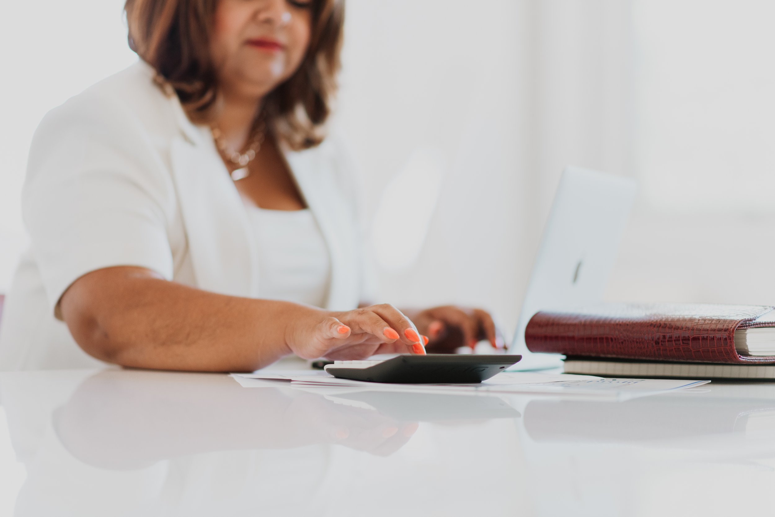 A woman with brown hair, wearing a white top, is sitting at a white desk using a tablet and a calculator. There is a large red book and papers on the desk.