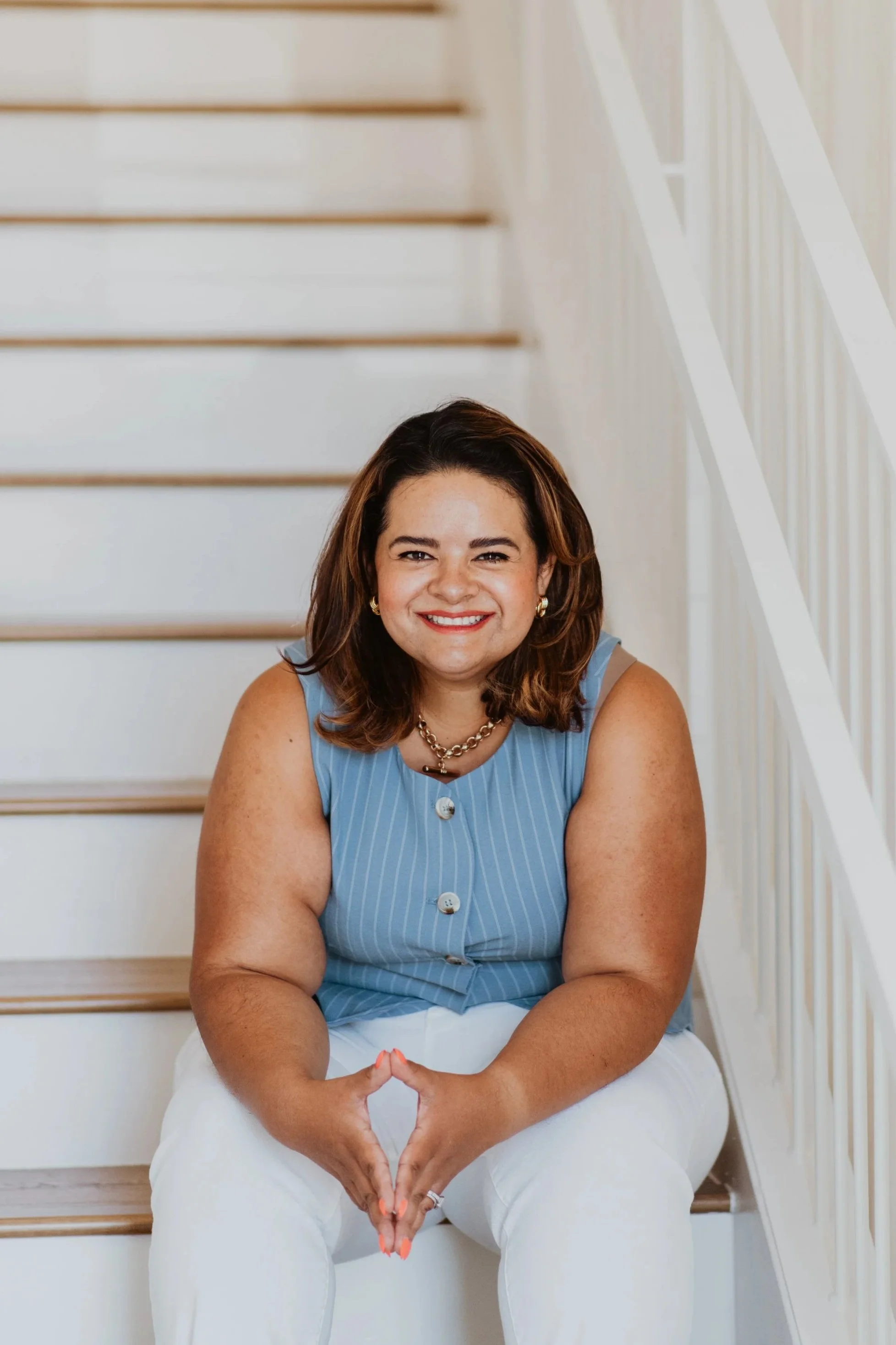 A woman with shoulder-length brown hair, wearing a light blue sleeveless top and white pants, sitting on stairs with a white railing, smiling at the camera, her hands forming a heart shape in front of her lap.