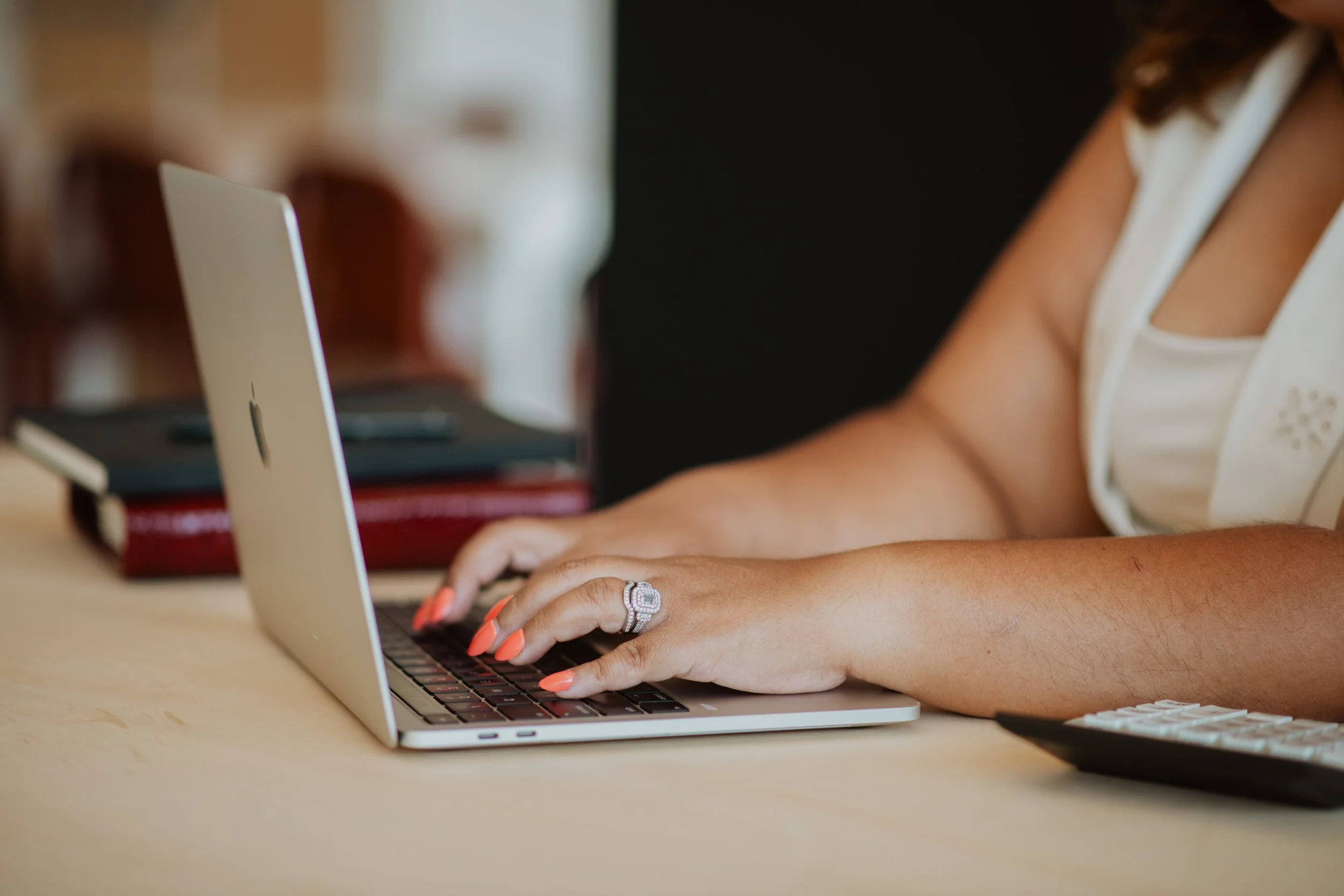 Close-up of a woman typing on a silver laptop, showing her right hand with an engagement ring and orange-painted nails. The background is blurred with some books and a dark surface.