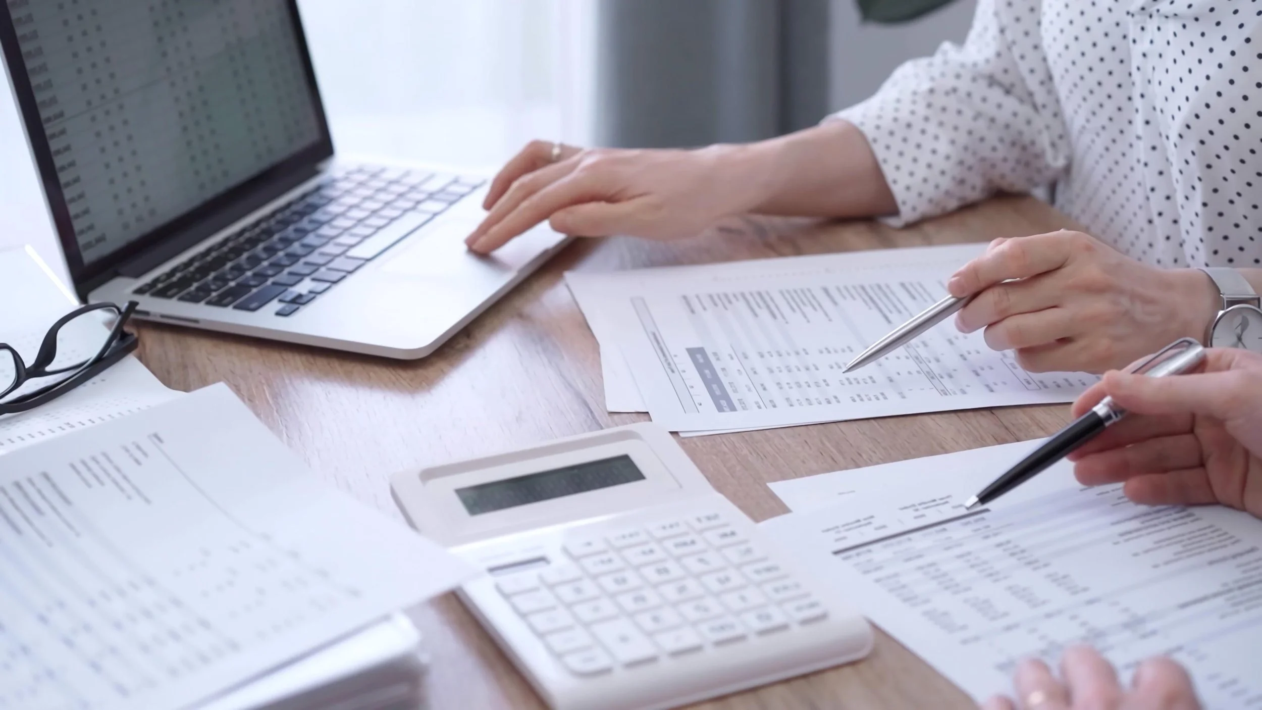 Person working with financial documents, a laptop, calculator, pen, and reading glasses on a wooden table.