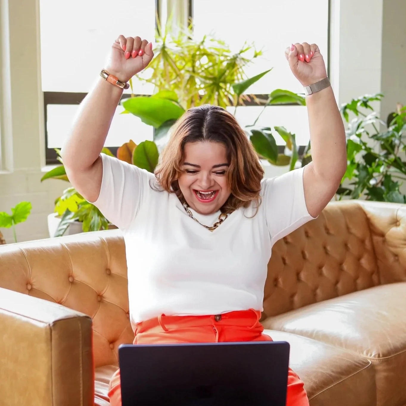 A woman sitting on a tan leather couch, smiling and raising her arms in celebration while looking at her laptop. She has short, wavy brown hair, is wearing a white t-shirt, orange pants, and jewelry. The background shows large green plants and a window.