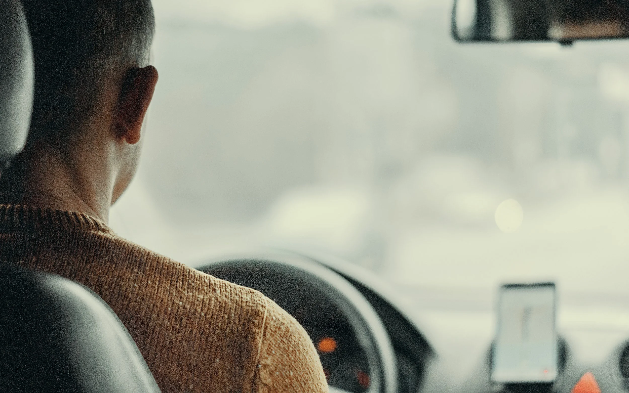 Rear view of a person with short hair in the driver's seat of a vehicle, wearing a mustard-colored sweater, looking out at a foggy or misty scene outside.
