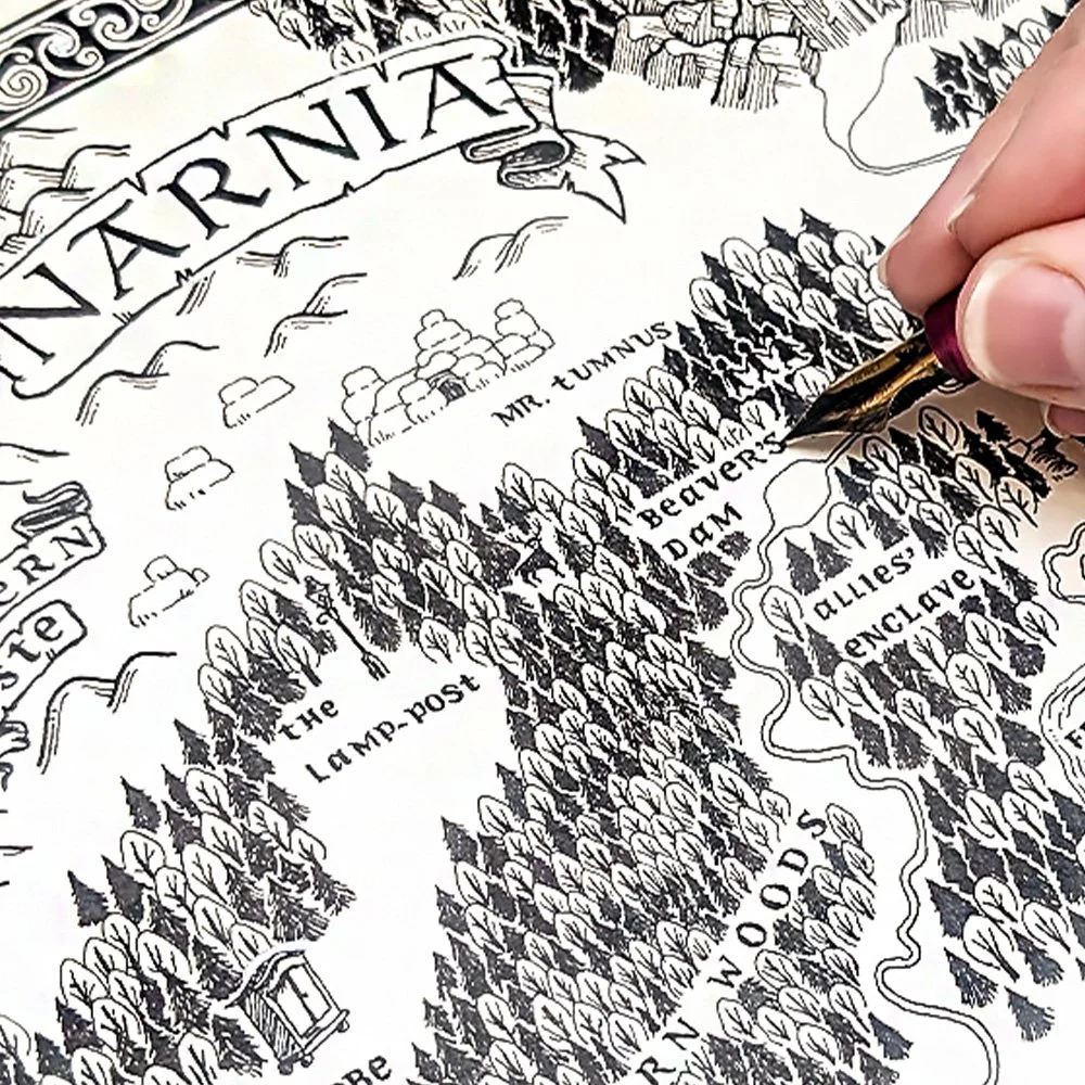 A person drawing a detailed map of Narnia with a fountain pen. The map shows towns, forests, and rivers.