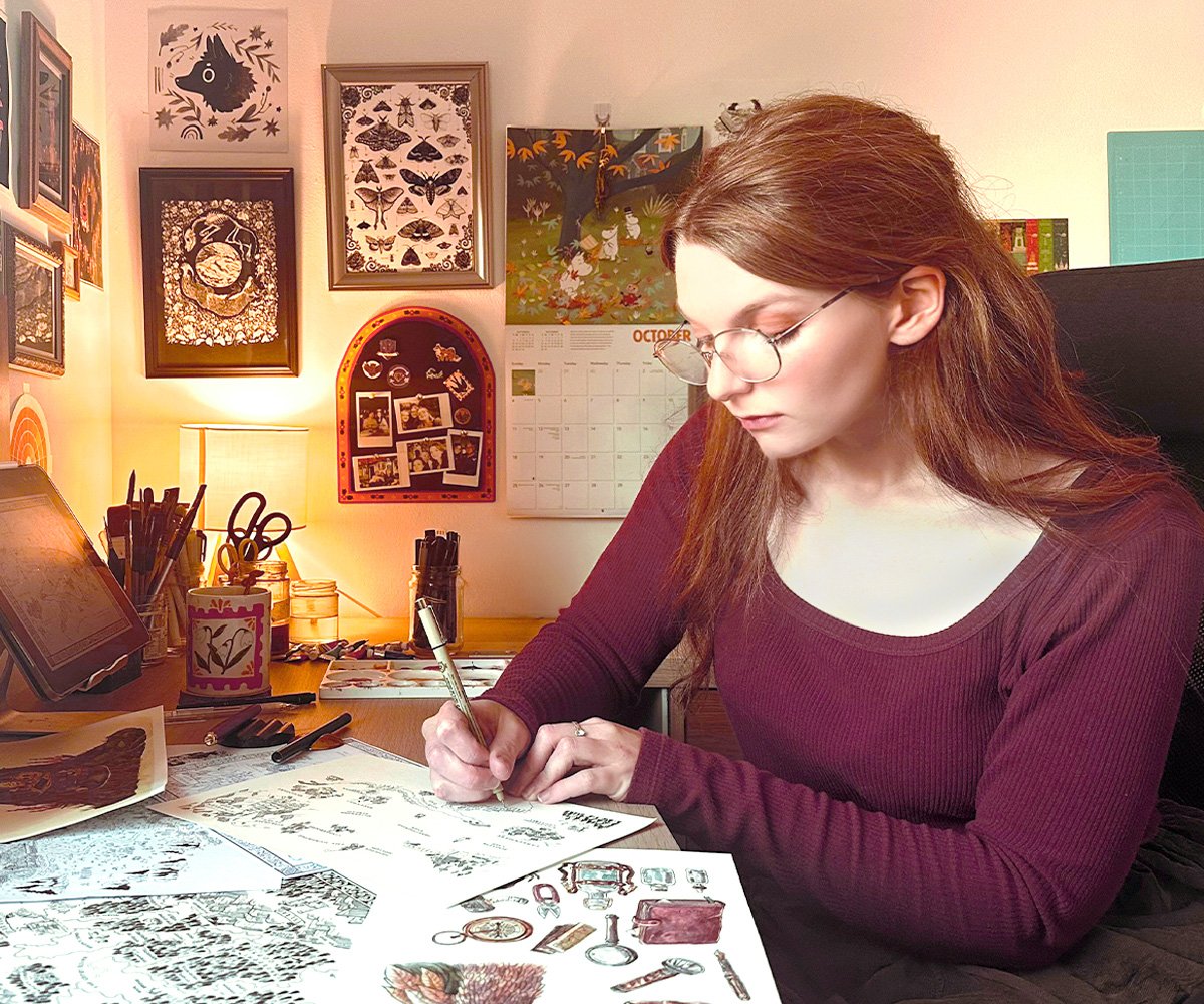 A young woman with glasses and long brown hair works on sketches at her desk. The desk has drawing supplies, photos, and artwork. The background shows framed art, a calendar, and a lamp.