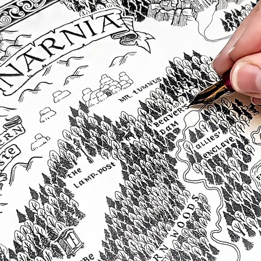 Close-up of a person drawing a detailed, black-and-white illustrated map with a fountain pen. The map features trees, mountains, and labeled locations such as 'The Lamp-post,' 'Mr. Tumnus,' and 'Narnia.'
