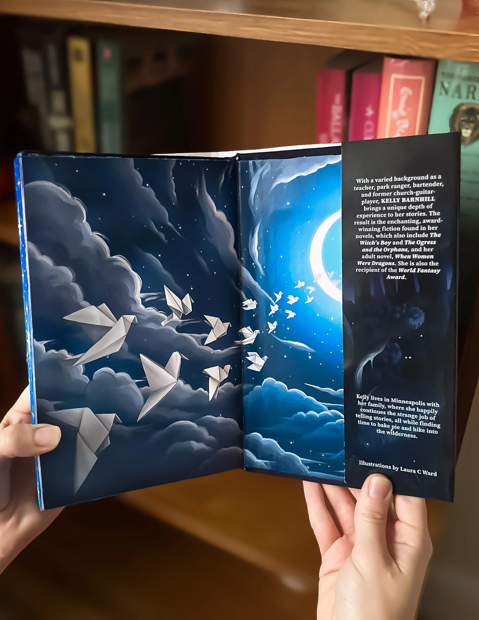 Open book with pages showing a dark sky scene with clouds, a crescent moon, stars, and paper birds flying through clouds. The right page contains text about author Kelly Barnhill.