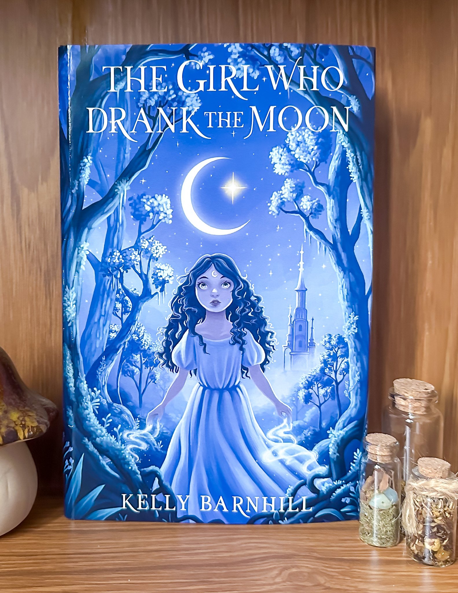 A book titled 'The Girl Who Drank the Moon' by Kelly Barnhill with a blue cover featuring a girl with long hair, a crescent moon, stars, and trees, placed on a wooden shelf with decorative bottles nearby.