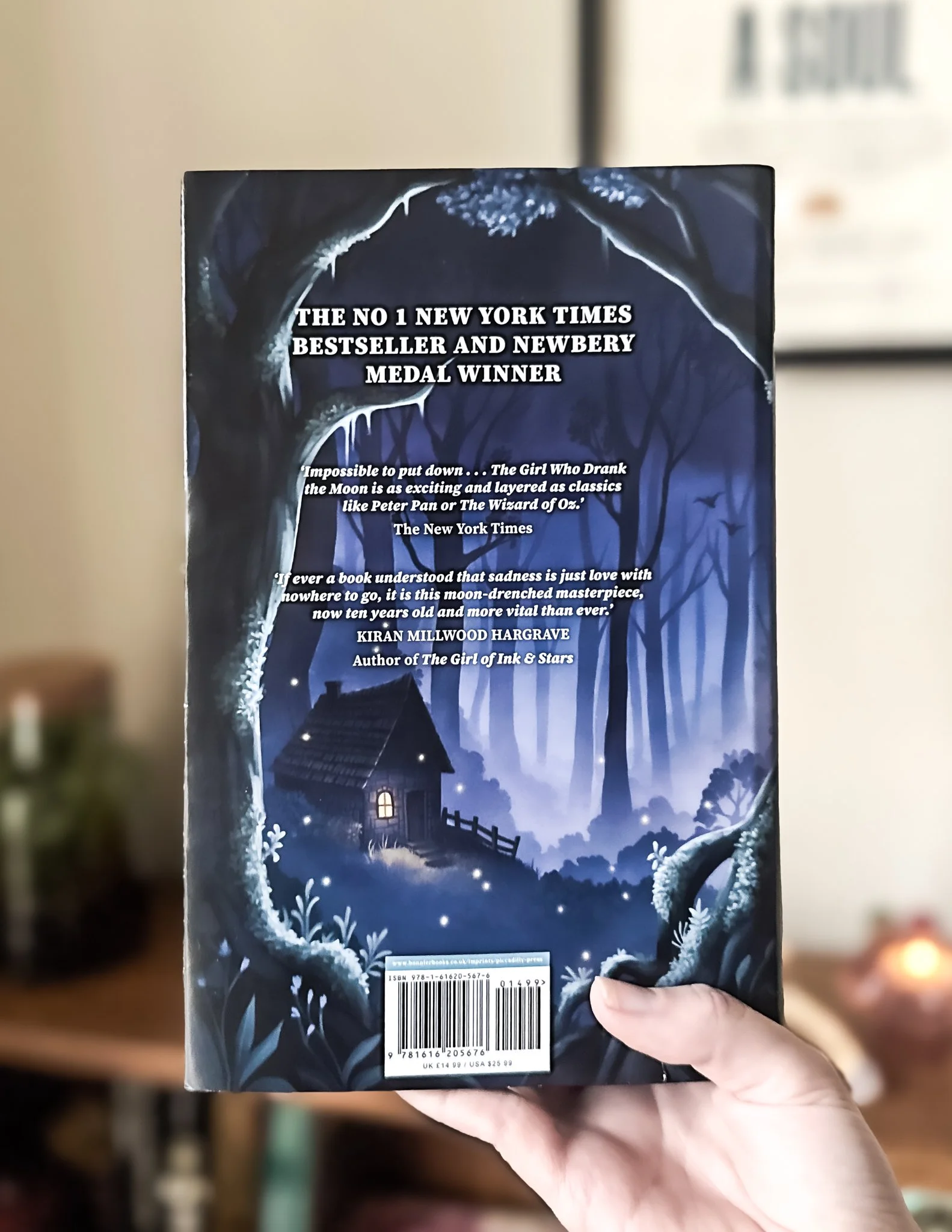 The back cover of a paperback book titled 'The Girl Who Drank the Moon' features a moonlit night scene with trees, a small house, and a fence, surrounded by a mysterious dark forest.