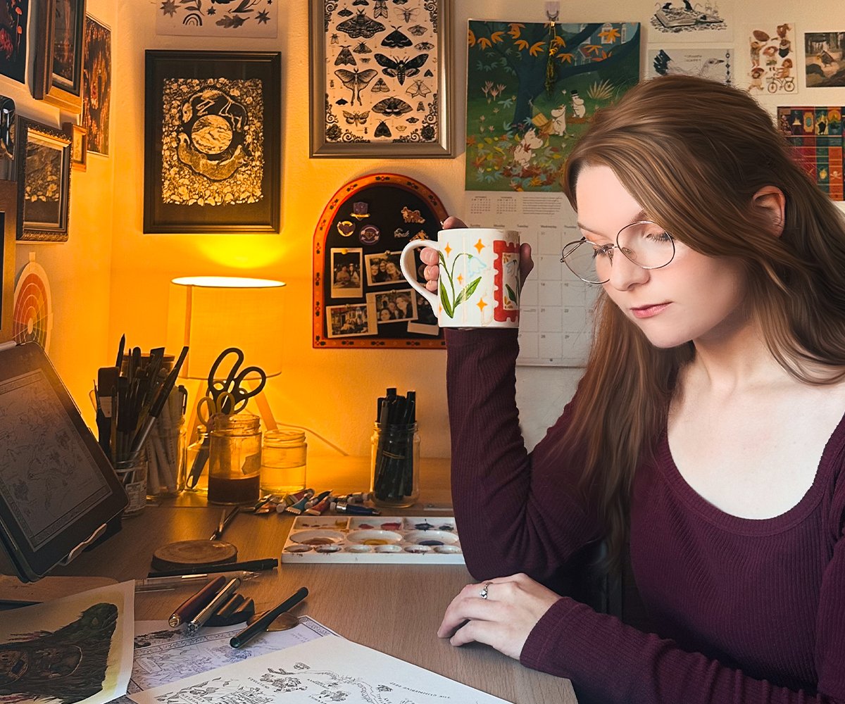 A young woman with long brown hair and glasses sits at an art workspace, holding a decorated mug. The desk has art supplies, including pens, watercolors, and sketches. The background features a warm yellow wall with framed artwork, photographs, and a lamp.