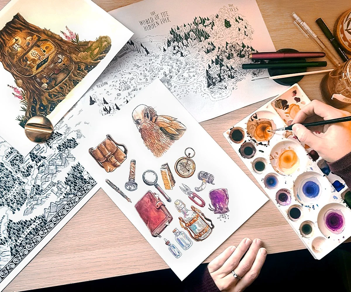 Overhead view of a table with art supplies and illustrations. There are color sketches, black-and-white drawings, and a watercolor palette. A person is painting with a brush, surrounded by markers, pens, and jars.