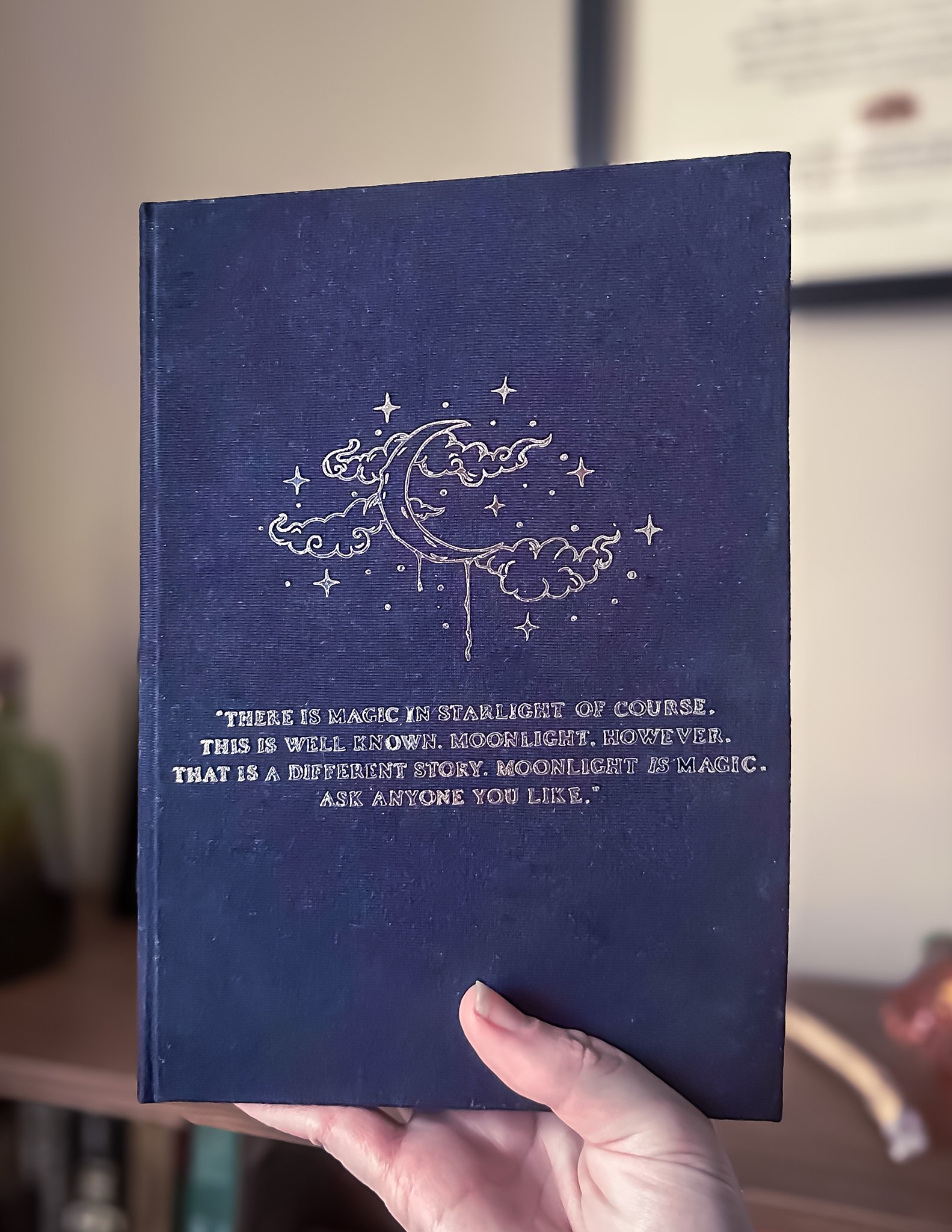 A person holding a dark blue book with an illustration of a crescent moon, clouds, and stars on the cover, and a quote about moonlight and magic.