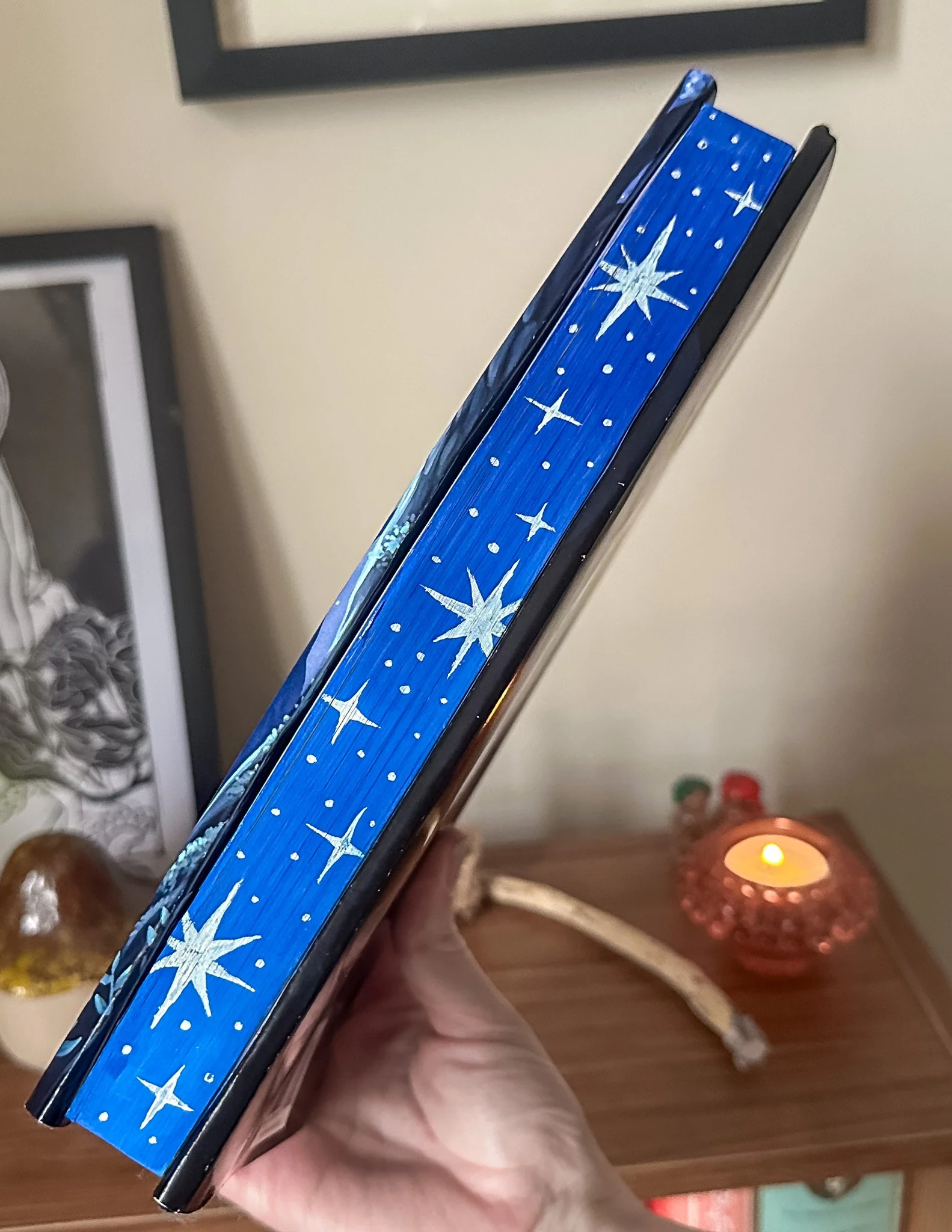 Close-up of a book's hand painted sprayed edges with a blue background and white star patterns being held in a person’s hand. In the background, a candle is lit on a wooden surface.