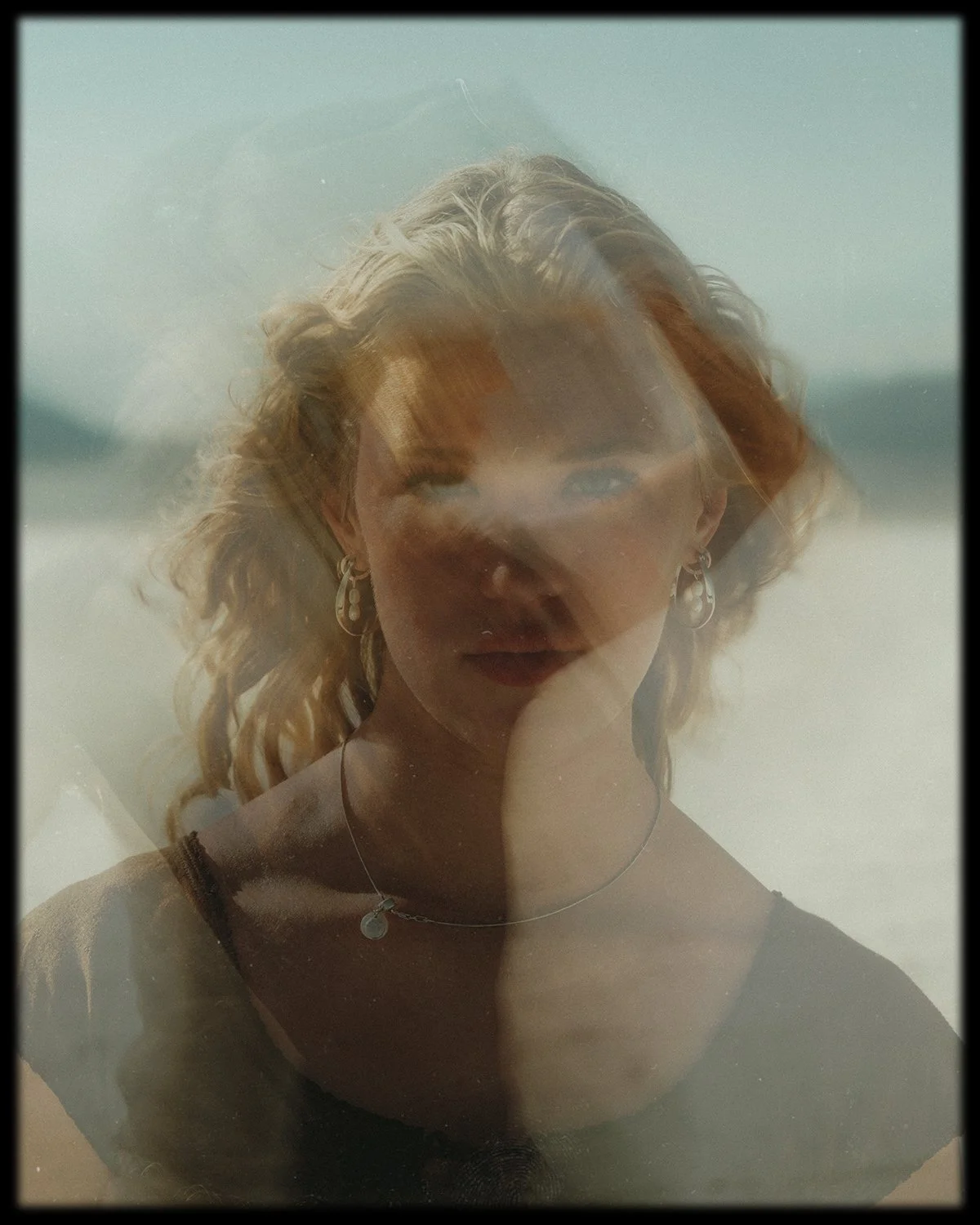 A reflection of a woman with blonde curly hair, wearing earrings and a necklace, seen through a glass window with a beach background.