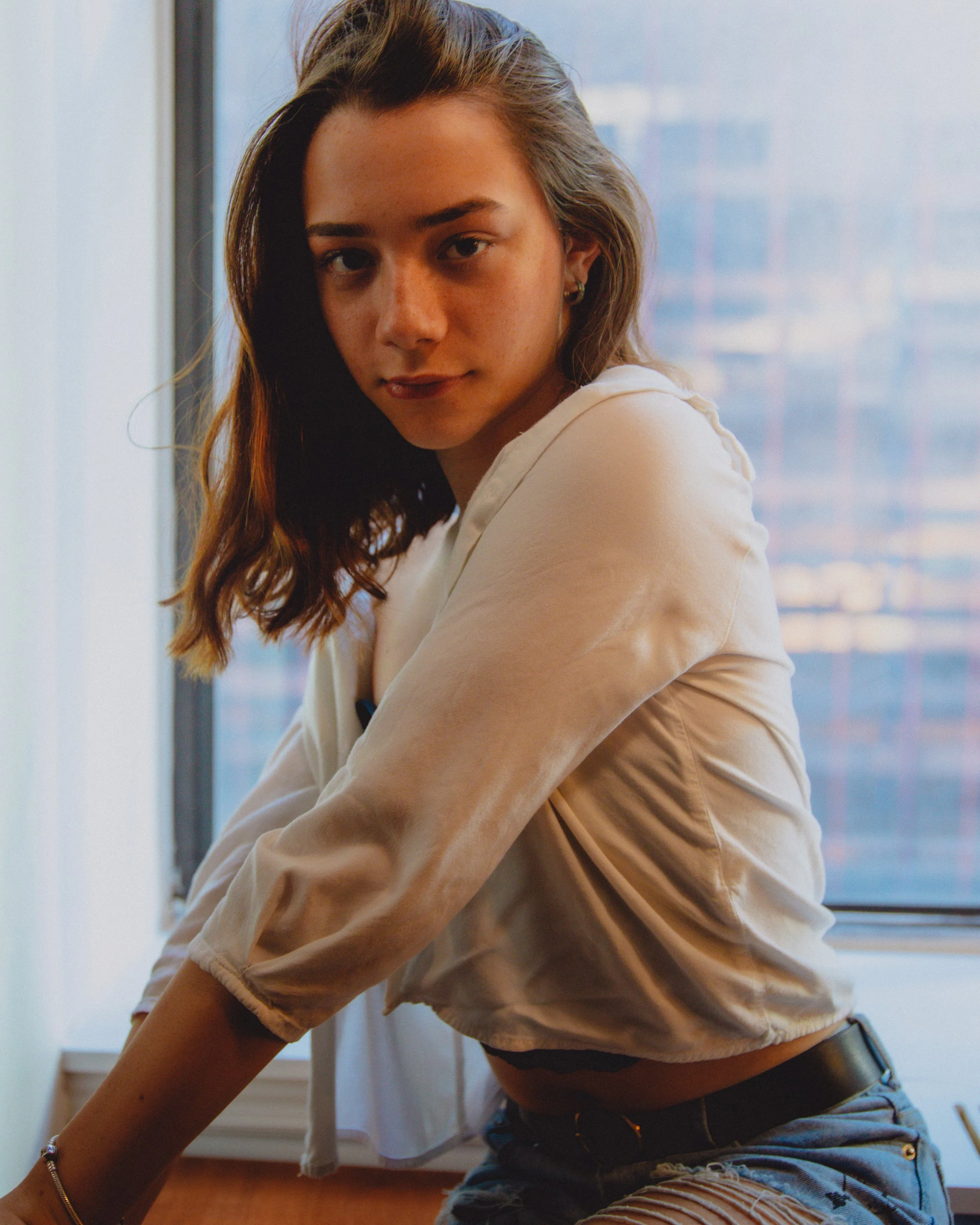 A young woman with light brown hair and a white shirt sitting near a window with buildings and a cloudy sky in the background.