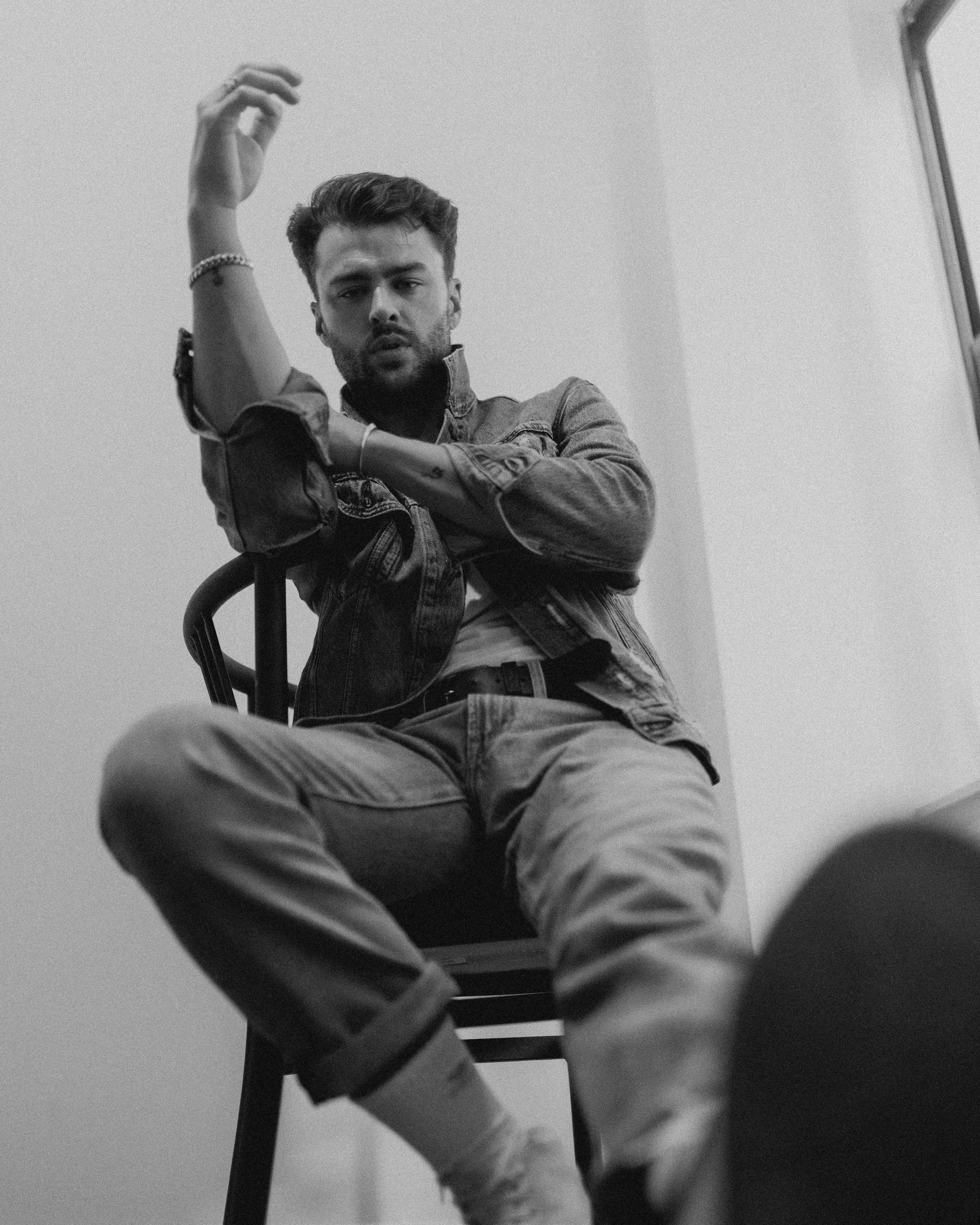 Black and white photo of a young man with styled hair, wearing a denim jacket and jewelry, sitting on a chair with legs stretched out, looking at the camera with a serious expression, in a plain indoor setting.