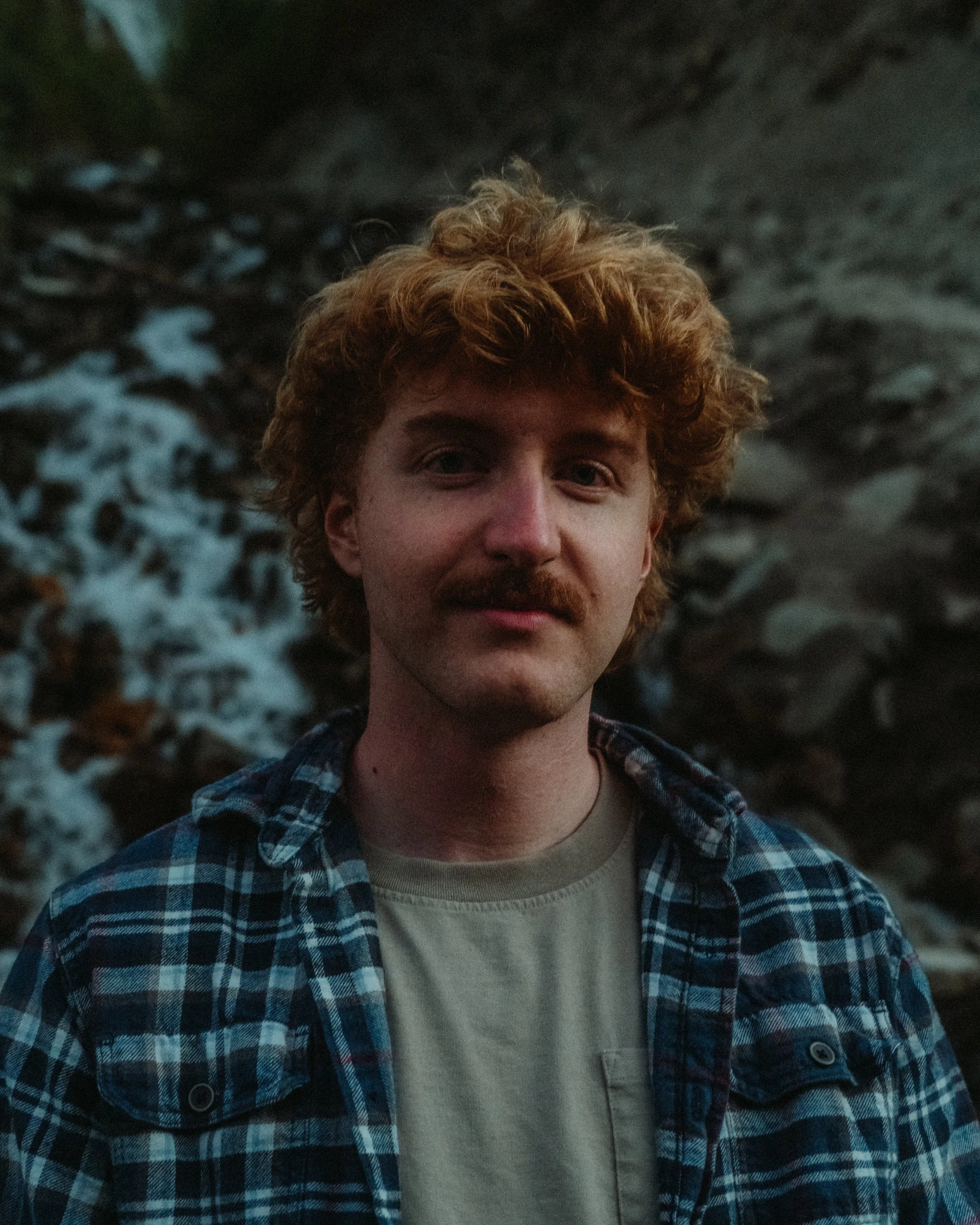 A young man with curly red hair, light facial hair, and a mustache stands outdoors near rocks. He is wearing a cream-colored shirt under a blue and black plaid jacket.