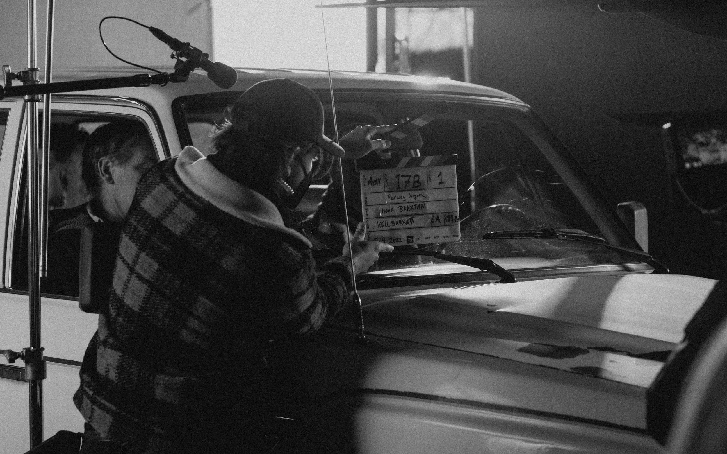 A person wearing a plaid jacket and cap holding a clapperboard while standing next to a car, with a person inside the car, on a film set.
